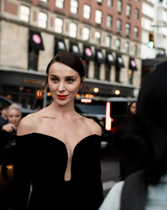 Woman in elegant black off-shoulder gown with red lips poses on a city street at night.