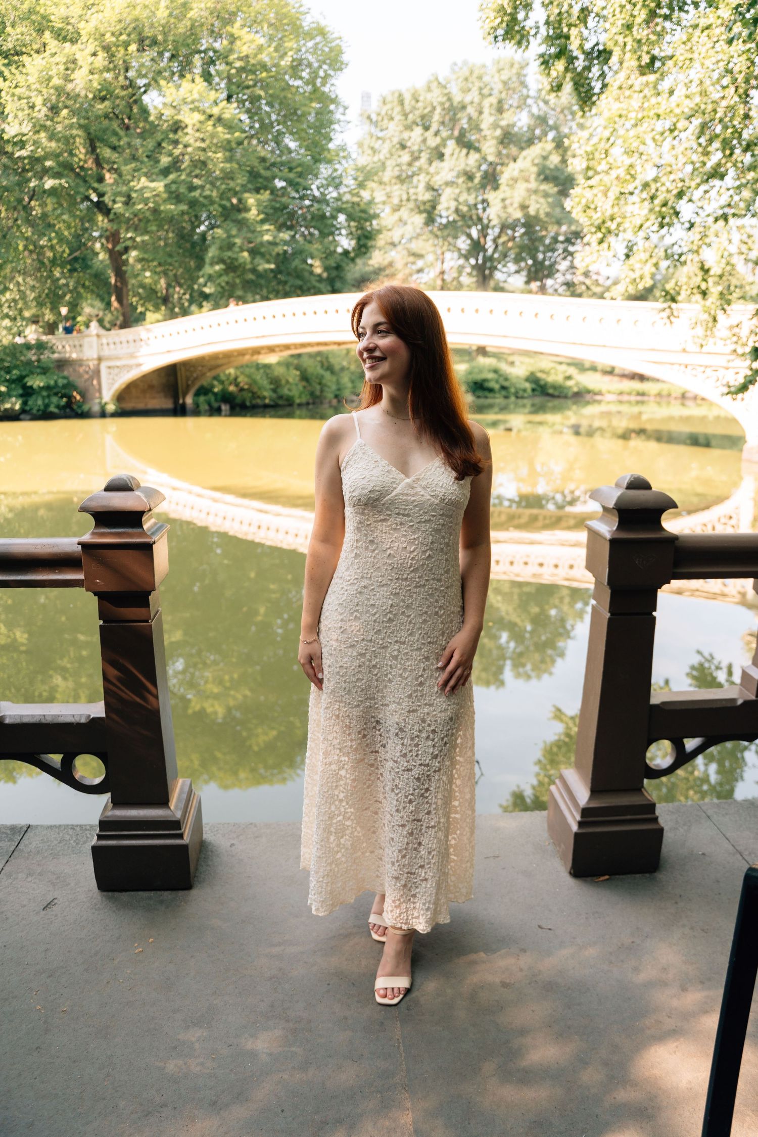 A woman in a white lace dress stands by a pond with a bridge in Central Park, New York.