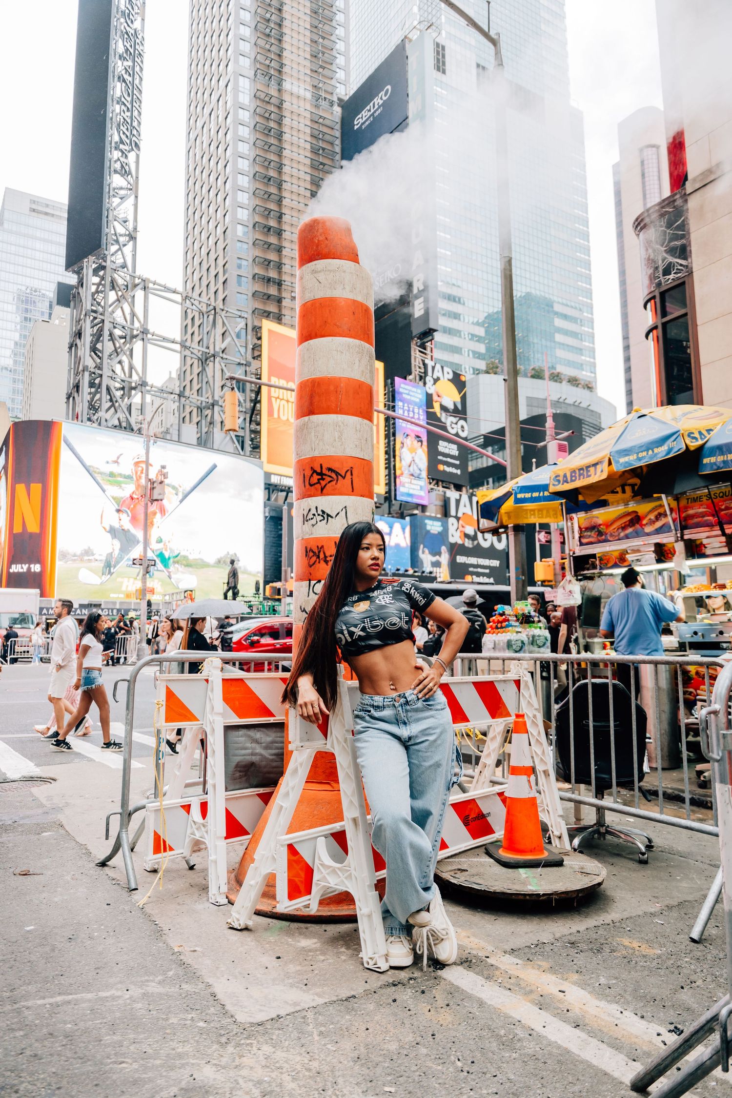 Woman in crop top and jeans posing at Times Square NYC construction site with orange barriers and steam pipe.