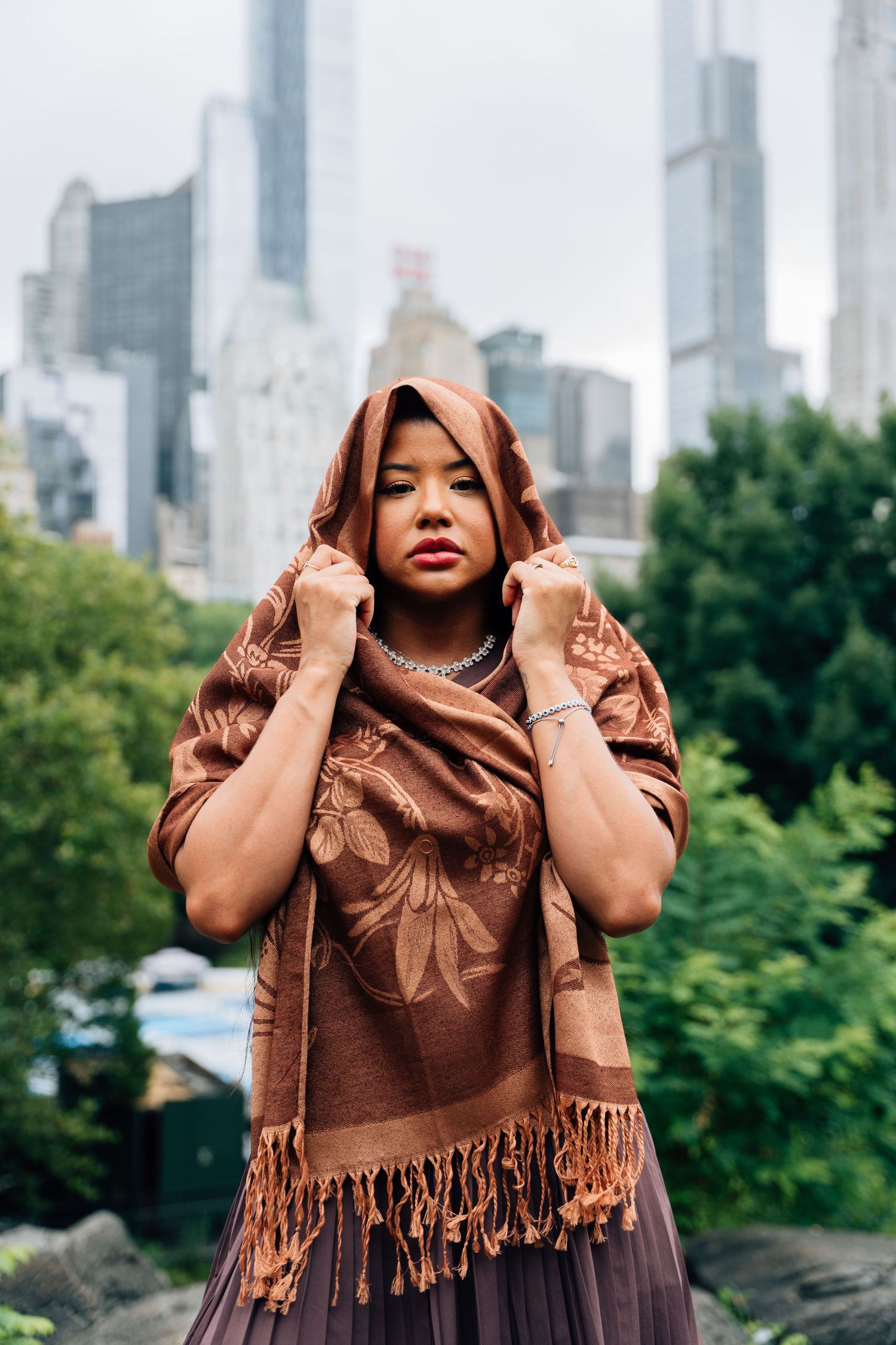 A young woman in a brown floral shawl draped over her head poses in Central Park with NYC skyline behind her.