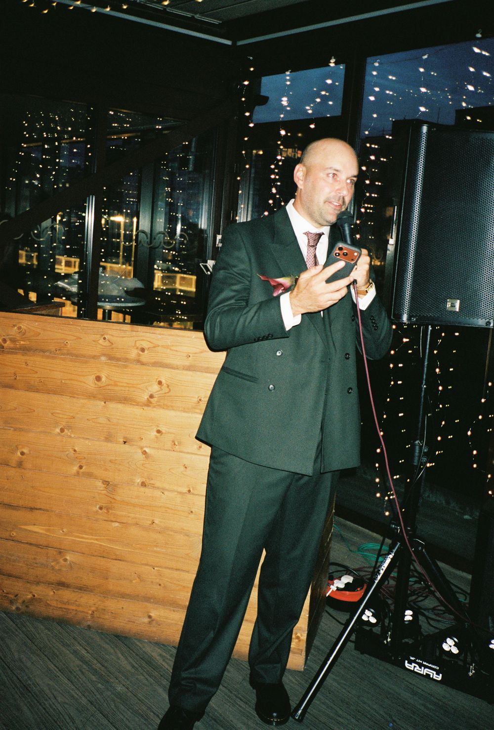 Bald man in dark suit speaking at microphone at elegant evening event with city lights in background.