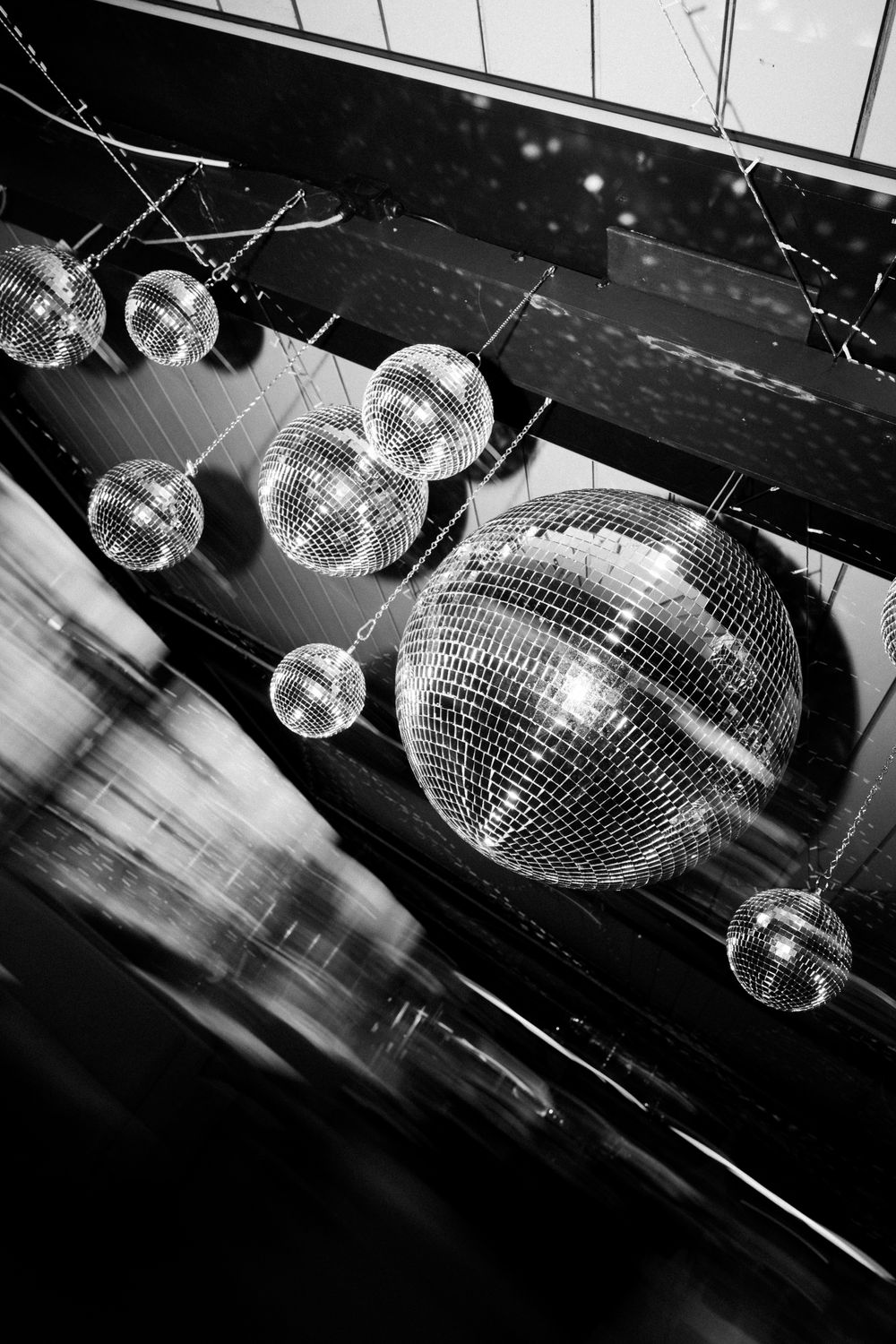 Black and white photo of multiple mirror disco balls hanging from ceiling, reflecting light in a dark venue.