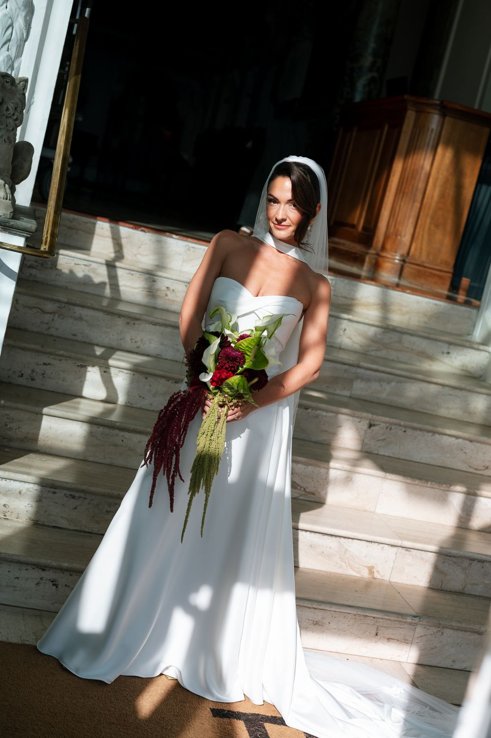 Bride in white strapless gown and veil holds red floral bouquet on marble steps in sunlight.