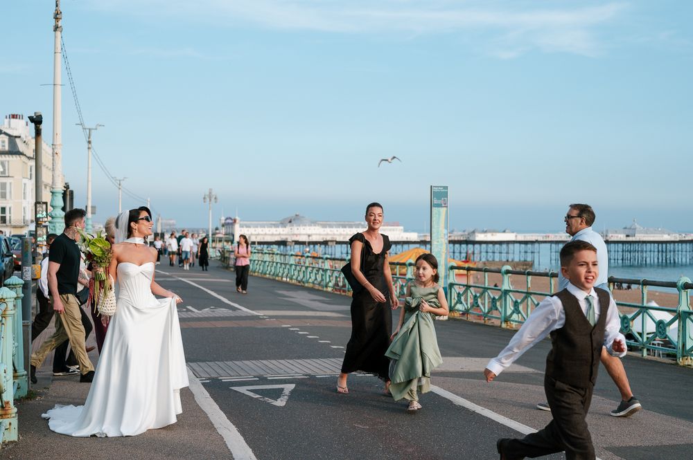 Wedding party walks on a seaside promenade with bride in white gown, bridesmaids, and children near Brighton Pier.