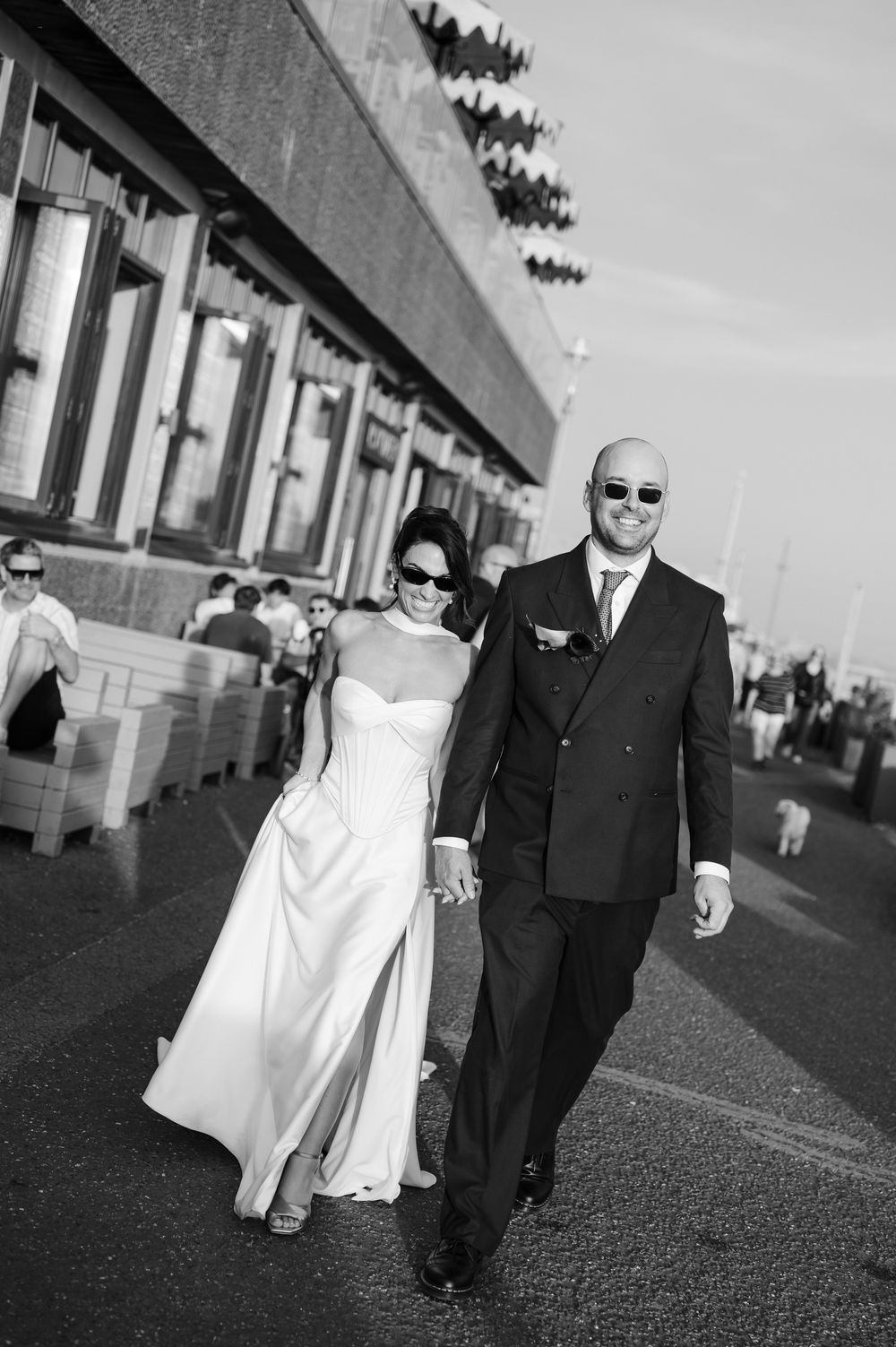 Black and white photo of a bride and groom holding hands, walking outside in wedding attire and sunglasses.