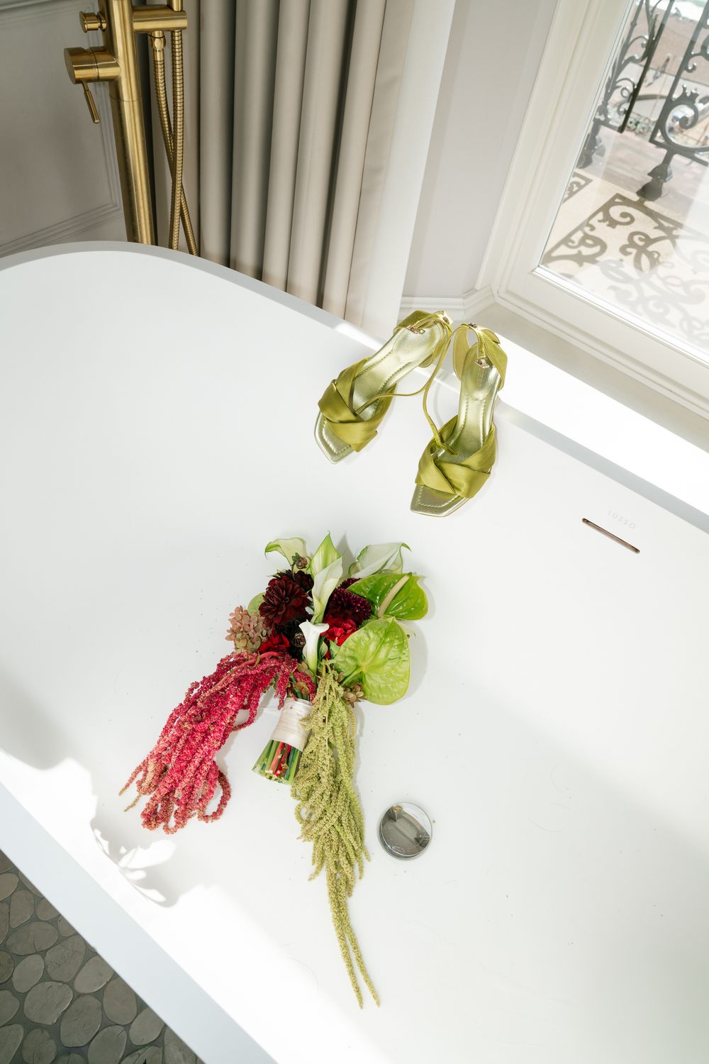 Gold strappy heels and a red floral bouquet displayed in a white freestanding bathtub near a bright window.