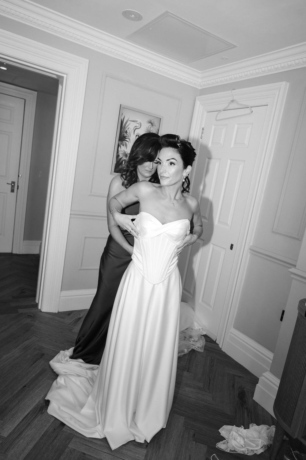 Bridesmaid helping bride in elegant white strapless wedding gown get ready in a classic hotel room.