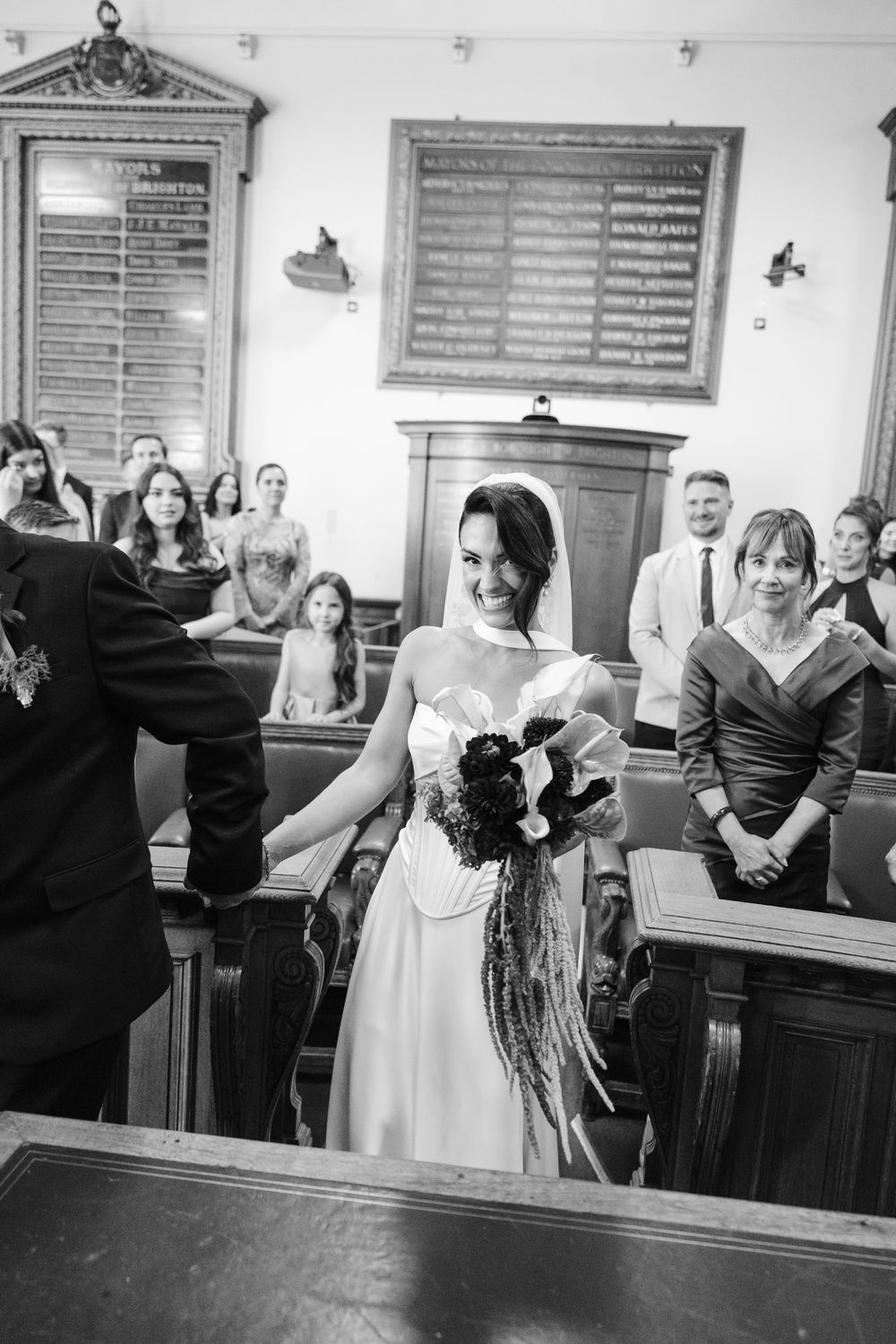 Bride smiling while walking down the aisle in a church wedding ceremony, holding a bouquet in black and white photo.