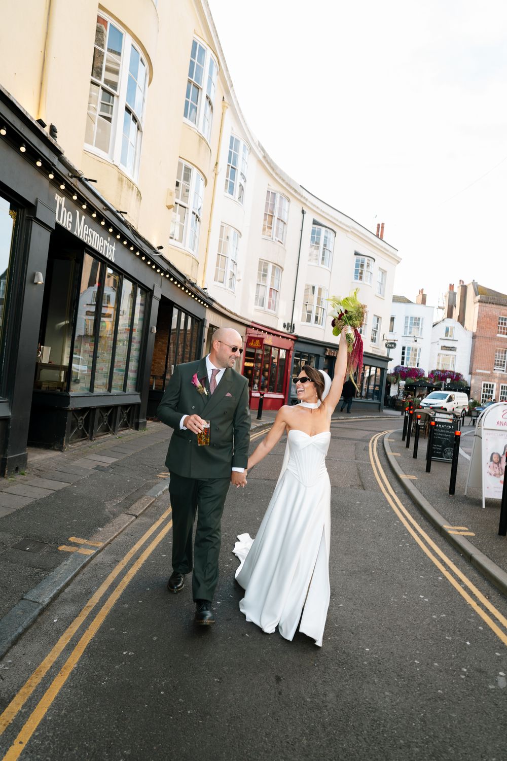 Bride and groom walking joyfully down a sunny UK street, bride raising bouquet in celebration on their wedding day.
