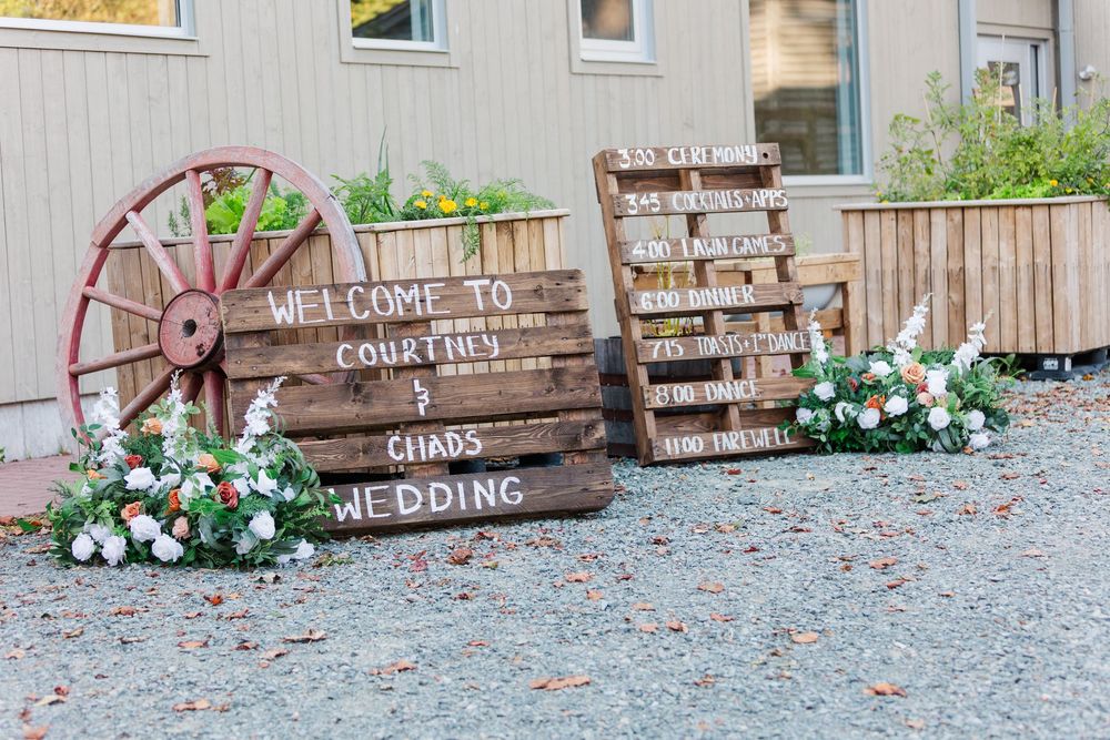 Rustic wooden pallet welcome sign for Courtney and Chad's wedding with a wagon wheel and white floral arrangements on a gravel path.