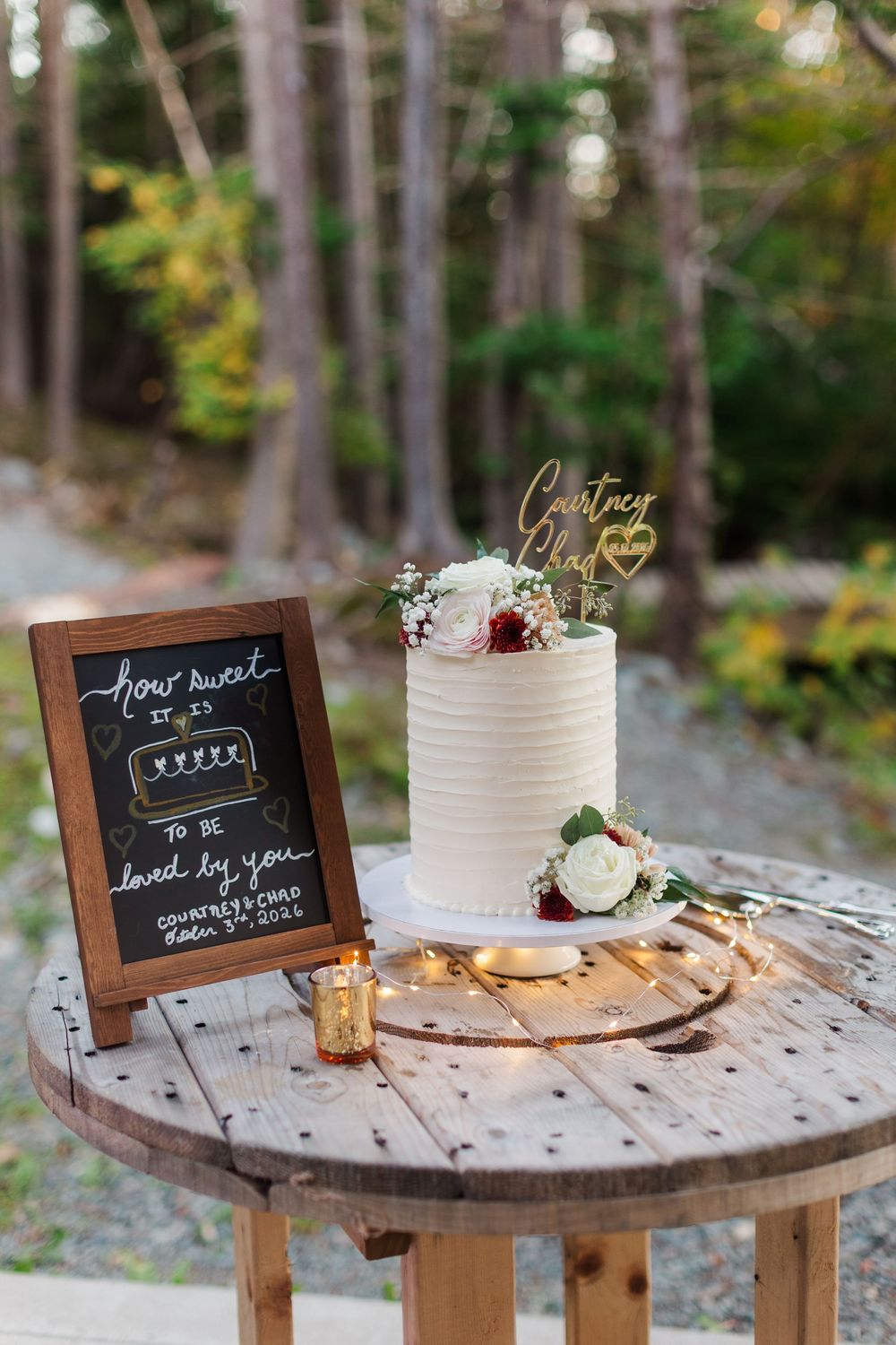 Rustic wedding cake with white buttercream and fresh flowers on a wooden spool table beside a chalkboard sign in a forest.