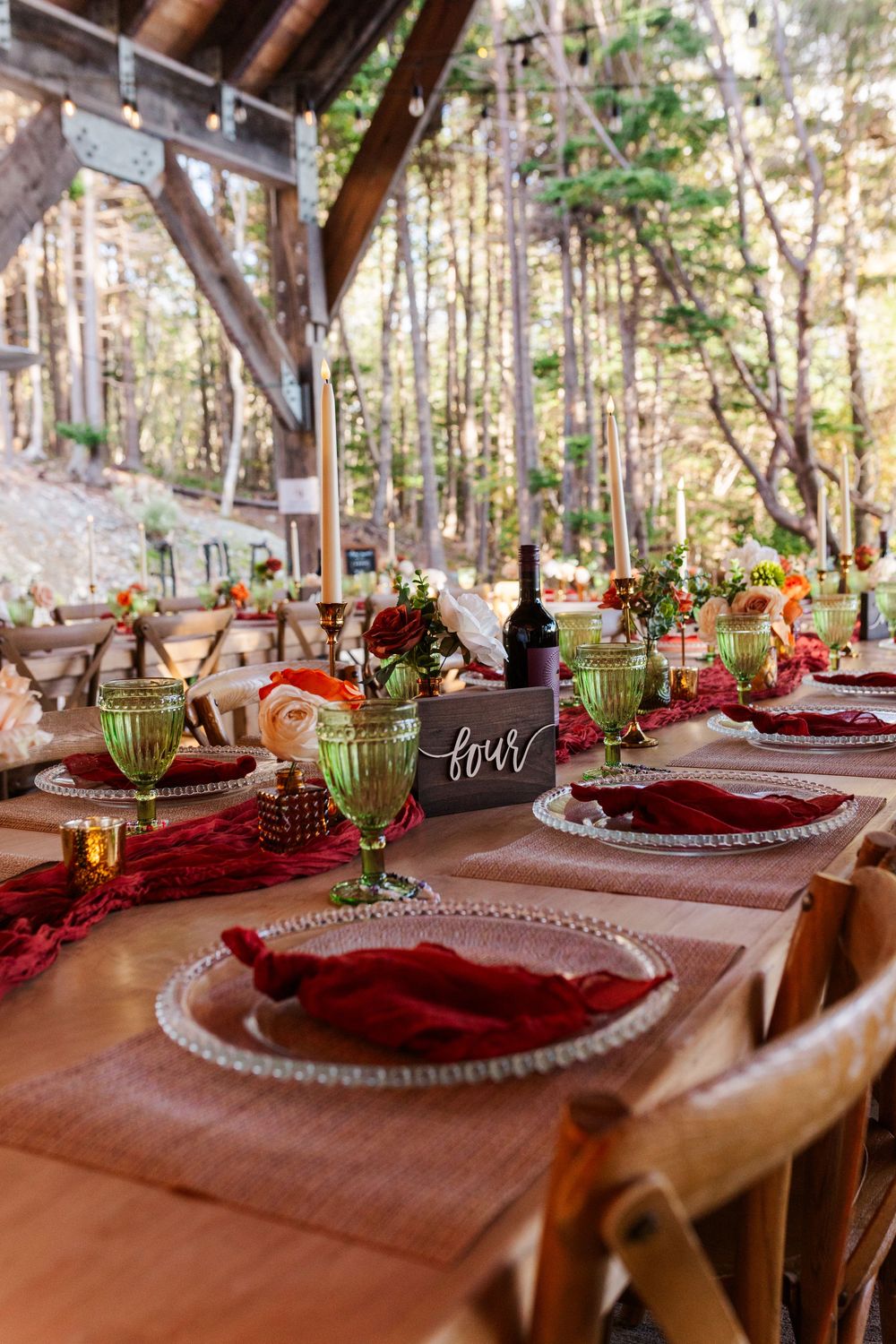 Rustic outdoor wedding table set with burgundy napkins, green goblets, beaded glass chargers, and floral centerpieces under a wooden pavilio