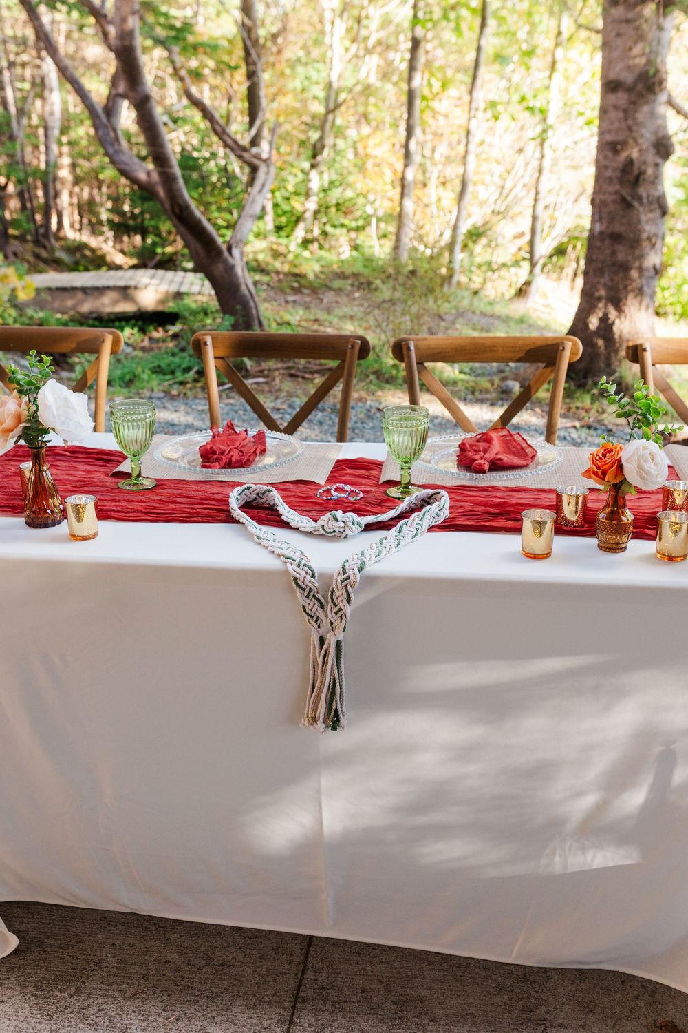 Outdoor wedding reception table with a rust red runner, green goblets, wooden chairs, gold votives, and a macrame handfasting cord.