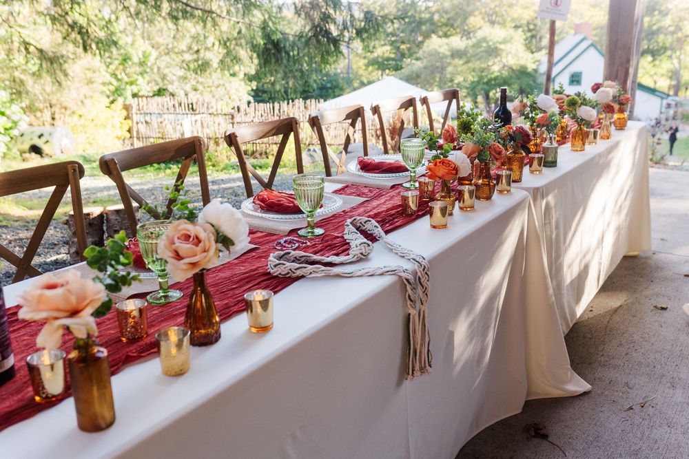Outdoor wedding reception table with white linens, red runner, peach roses in amber glass vases, candles and wooden chairs.