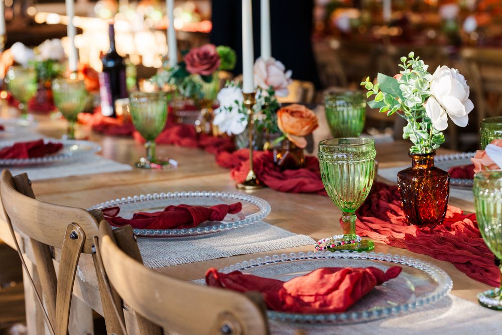 Elegant wedding table setting with red napkins, green glass goblets, white floral centerpieces, candles and rustic wooden chairs.