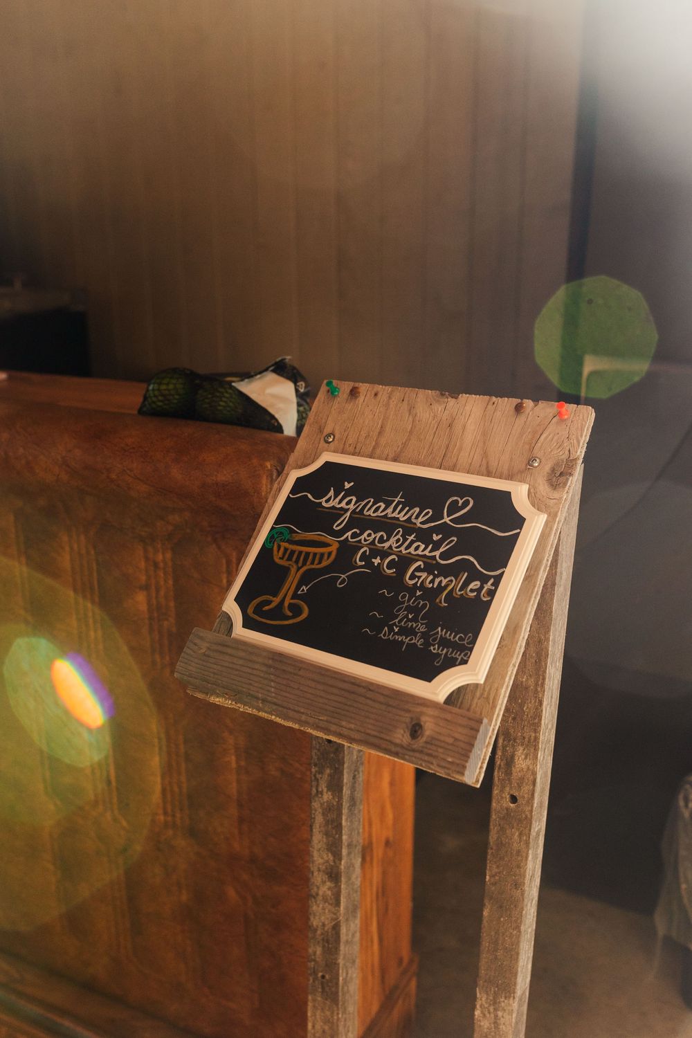 Rustic wooden easel holding a chalkboard sign for a signature cocktail, the C+C Gimlet, displayed beside a weathered wood bar.