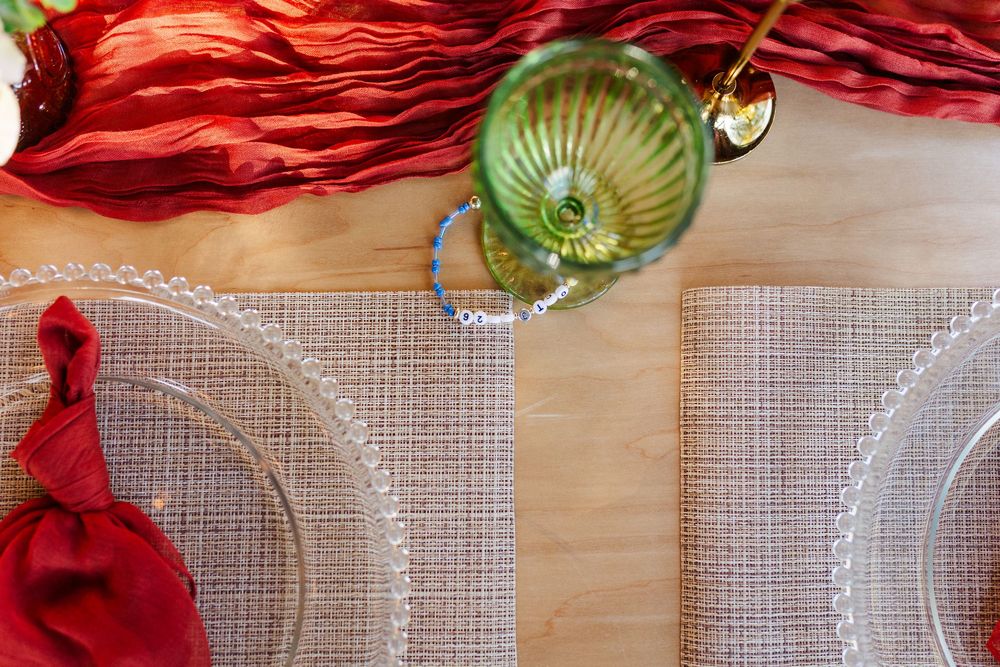 Elegant table setting with a green vintage goblet, beaded glass charger plates, woven placemats and a red napkin on a wooden table.