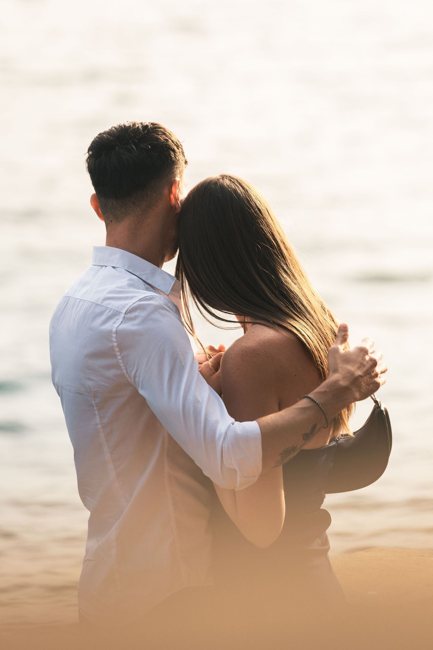 Couple embracing from behind on a sunlit beach at sunset, the man kissing the woman's head as the calm sea glows behind them.