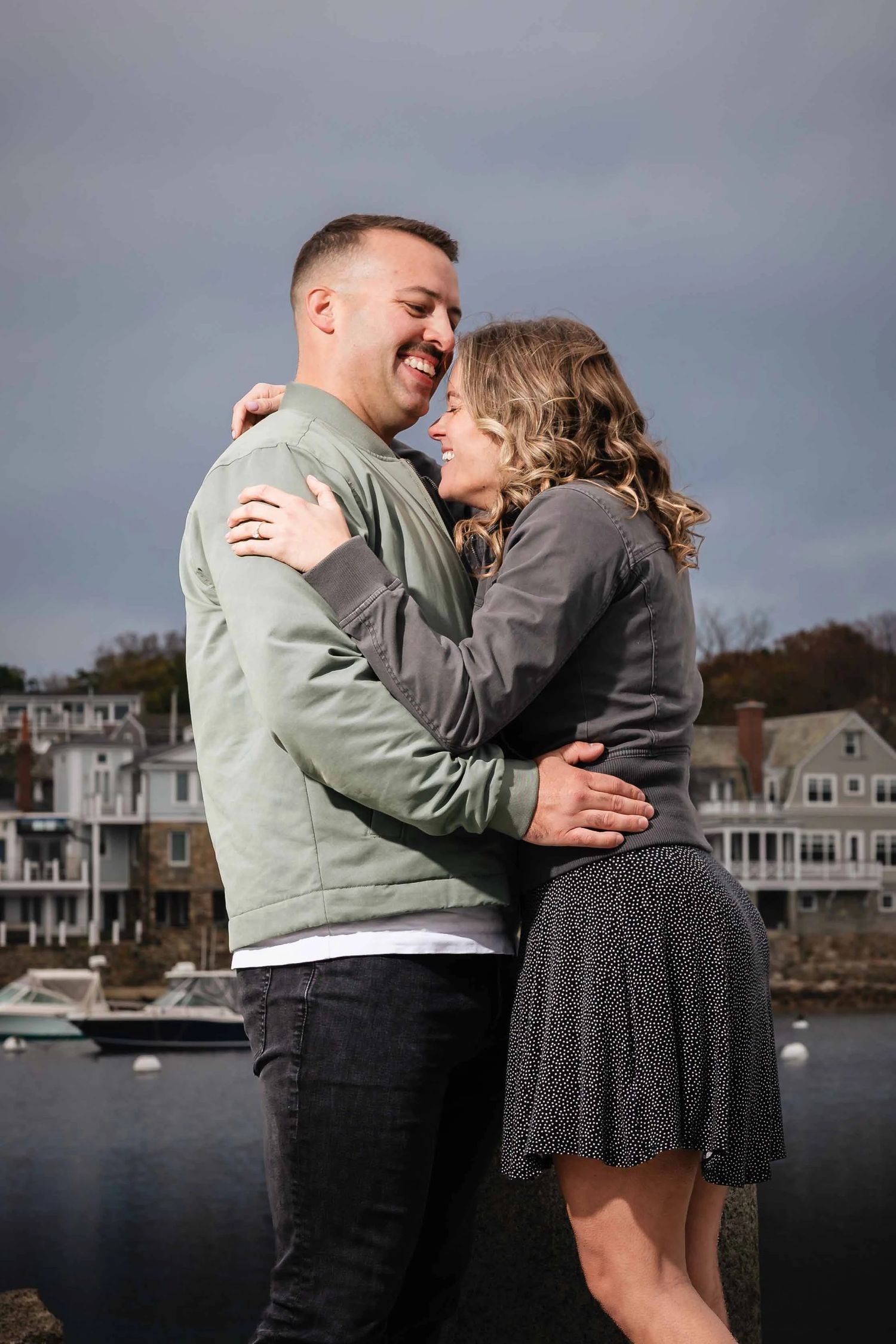 Man and woman embracing and laughing together outdoors by the water. Engagement at Rockport Harbor, Rockport, MA