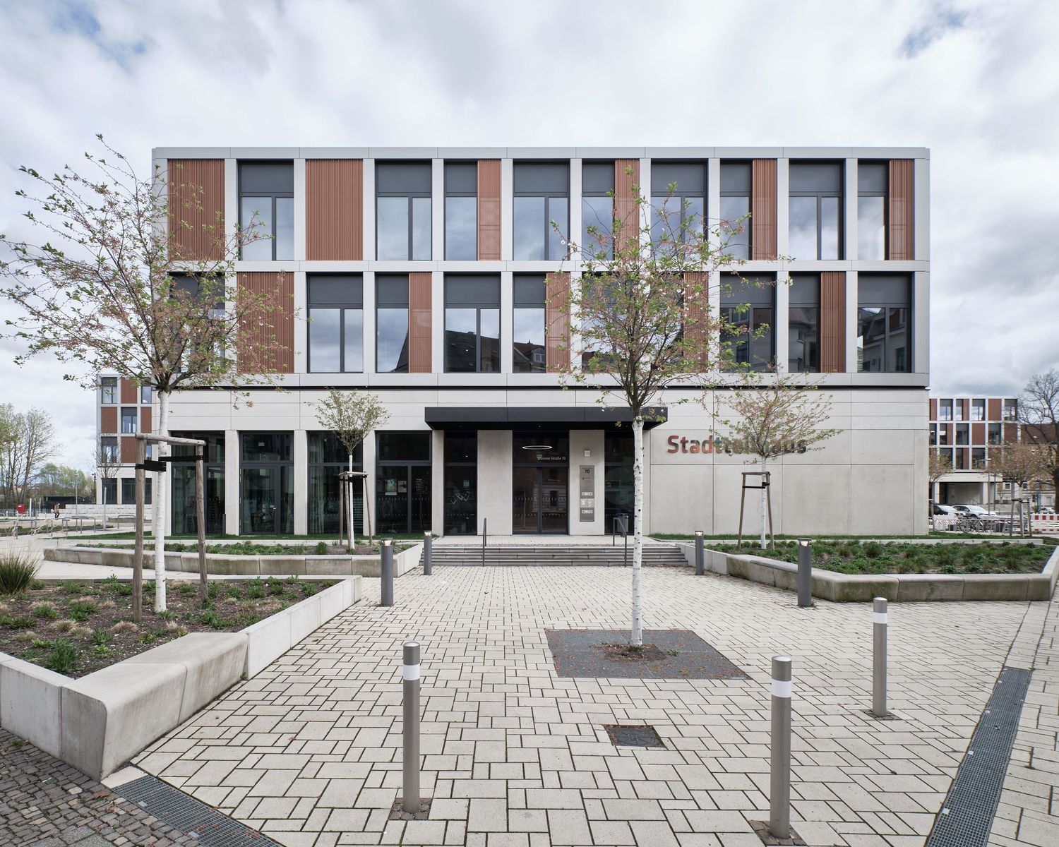 Modern multi-story Stadthaus building with brick and glass facade, paved plaza, young trees and landscaped planters on a cloudy day.
