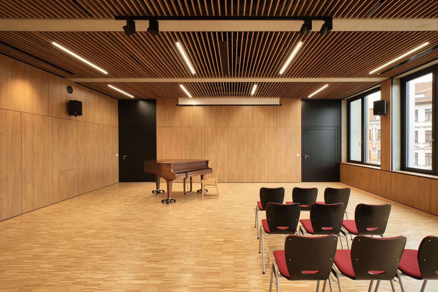 Modern wood-paneled recital hall with a grand piano, red chairs, parquet flooring, slatted ceiling lights and large windows.