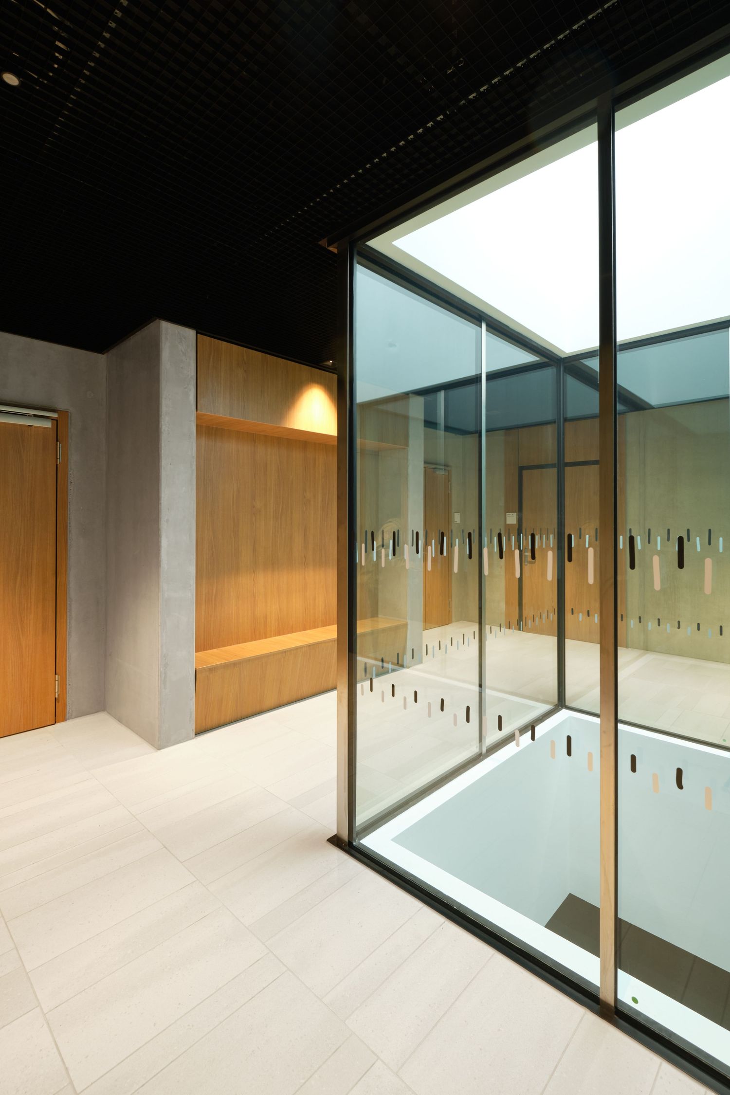 Modern interior with a glass-enclosed light well, warm wood paneling, a built-in bench, and pale tile flooring beneath a dark ceiling.