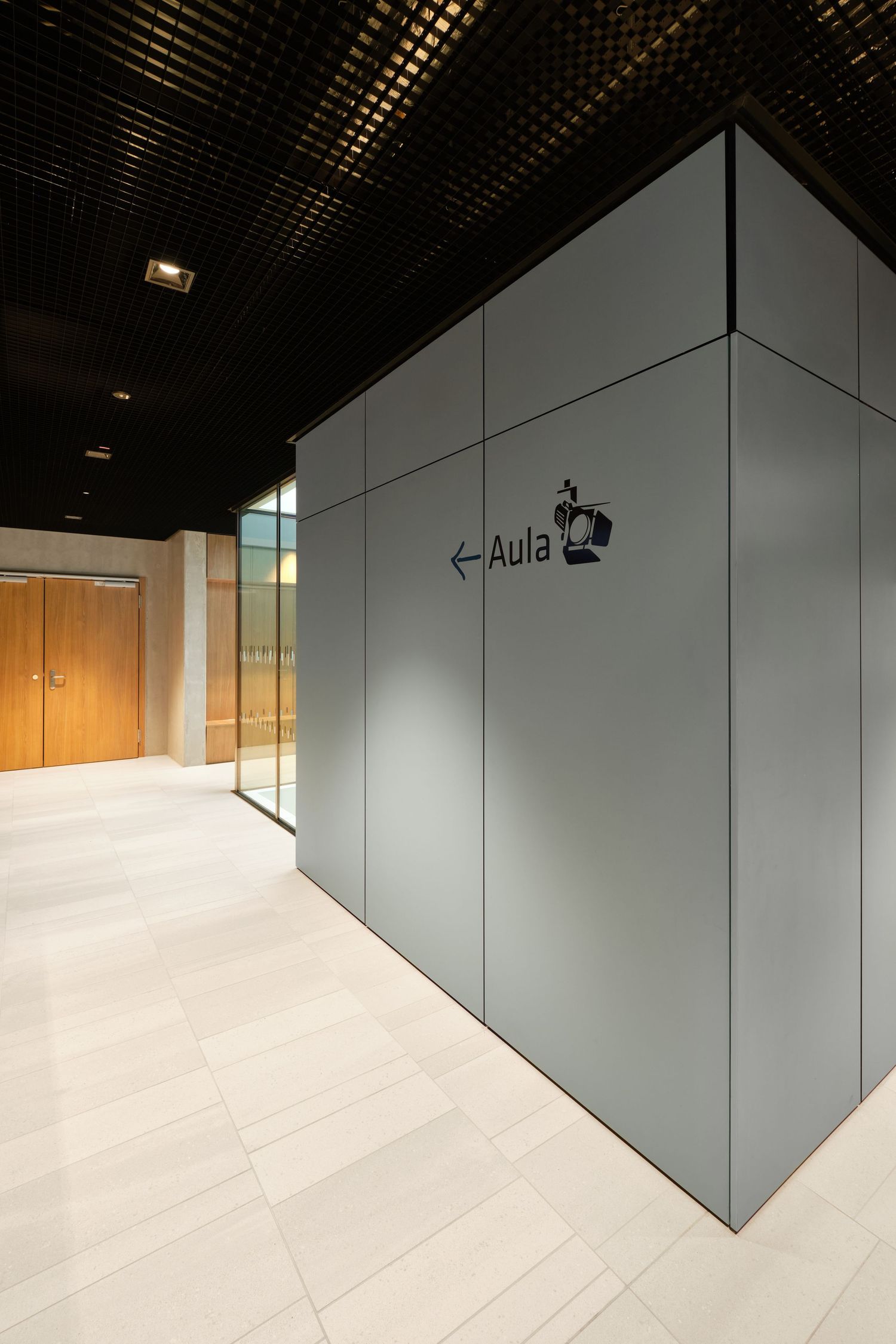 Modern corridor with gray wall panels and an Aula directional sign with arrow, light tile floor, dark grid ceiling and wooden doors.