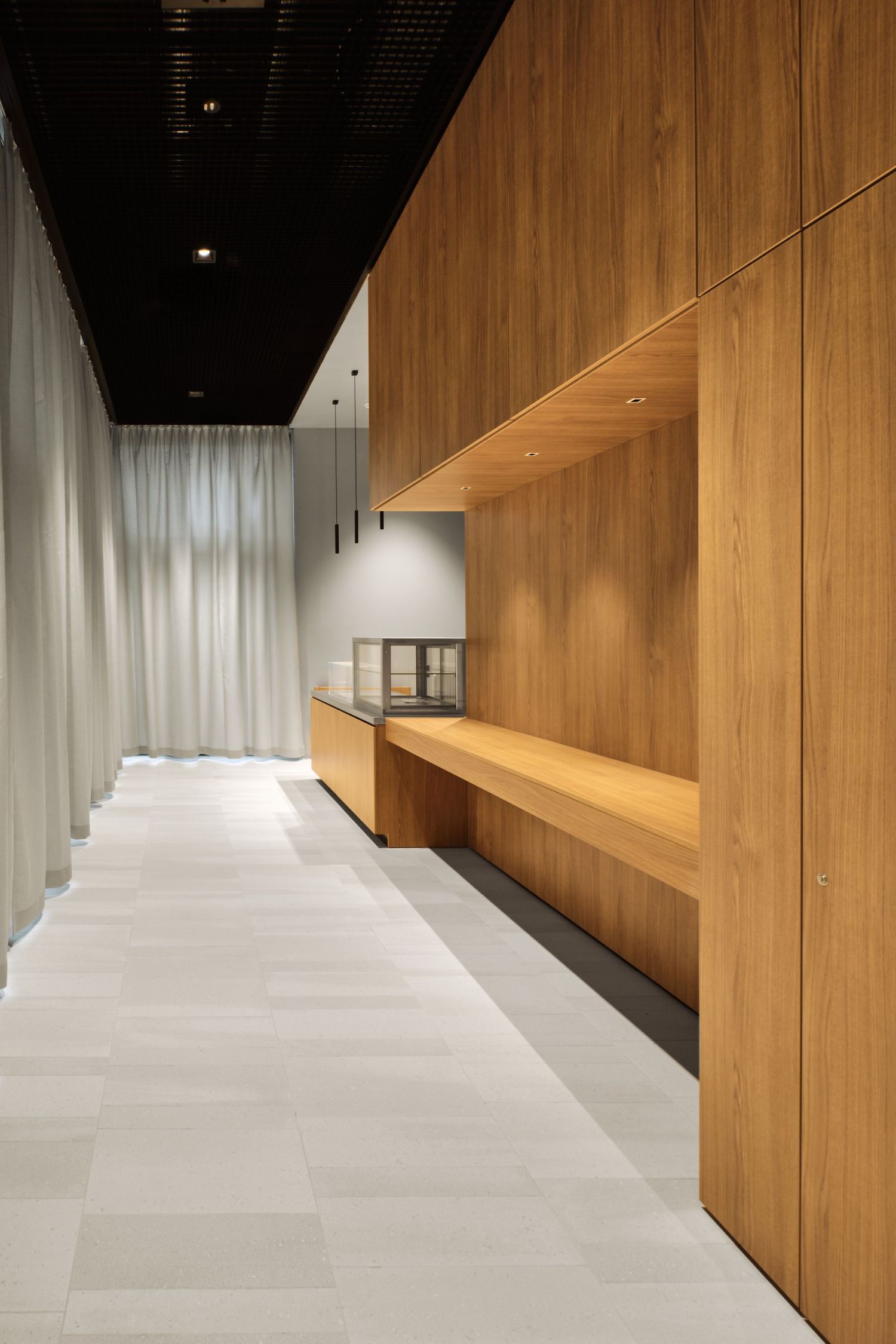 Modern minimalist corridor with warm wood paneling, built-in oak bench, glass display case, sheer curtains and light stone tile flooring.