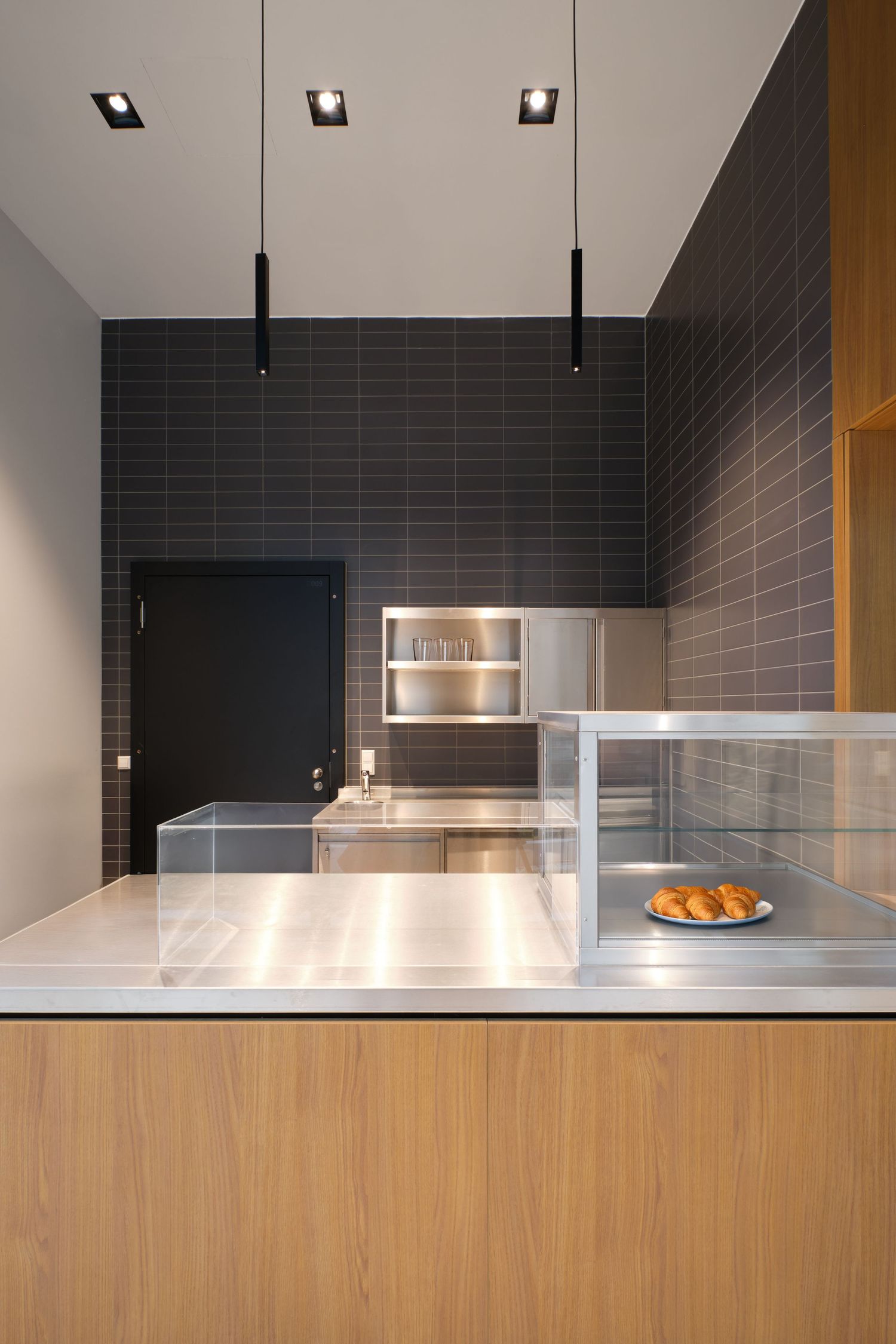 Modern café counter with stainless steel top, oak front, glass display case with croissants, dark tiled walls and black pendant lights.
