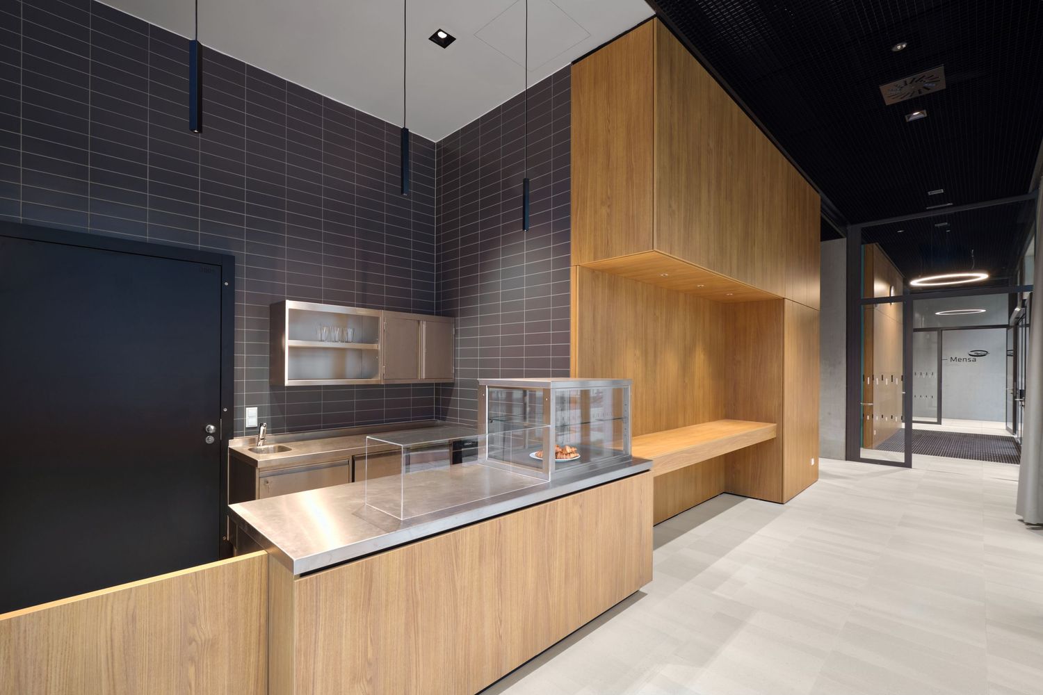 Modern cafe counter with light wood paneling, stainless steel prep surface, glass display case and dark tiled walls in a sleek interior.