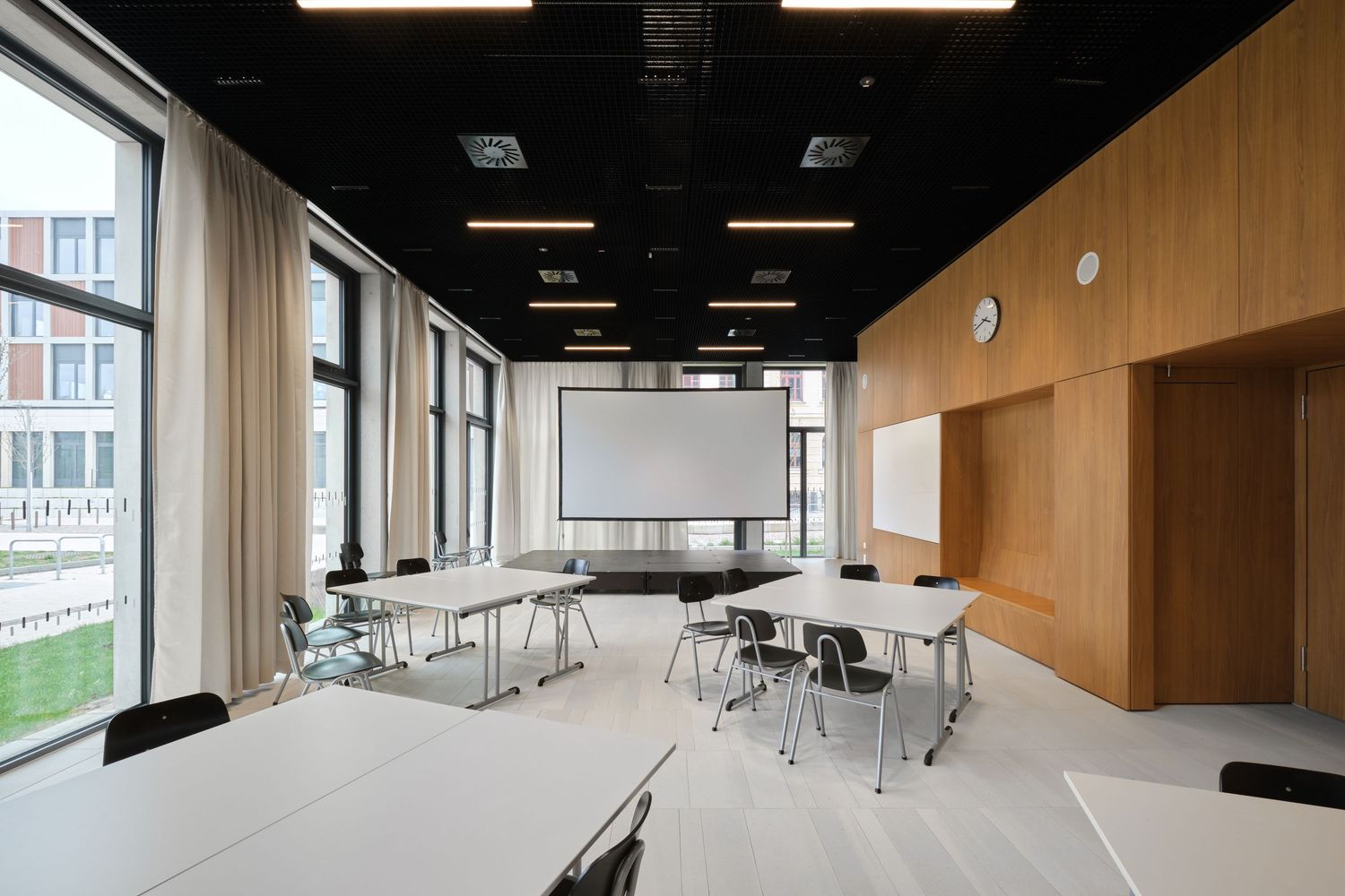 Bright modern meeting room with wood paneled walls, tables and chairs, large windows and a projector screen on the wall.