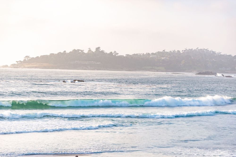 Turquoise ocean waves roll toward a sandy beach in soft morning haze, with a tree-covered rocky headland on the horizon.