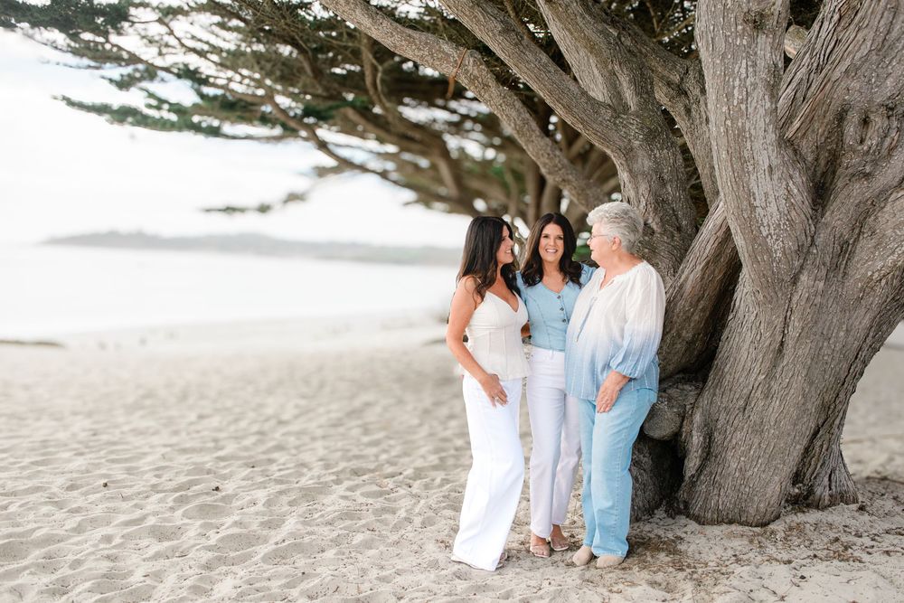 Three women in white and soft blue outfits stand together beside a weathered cypress tree on a sandy beach near calm ocean water.