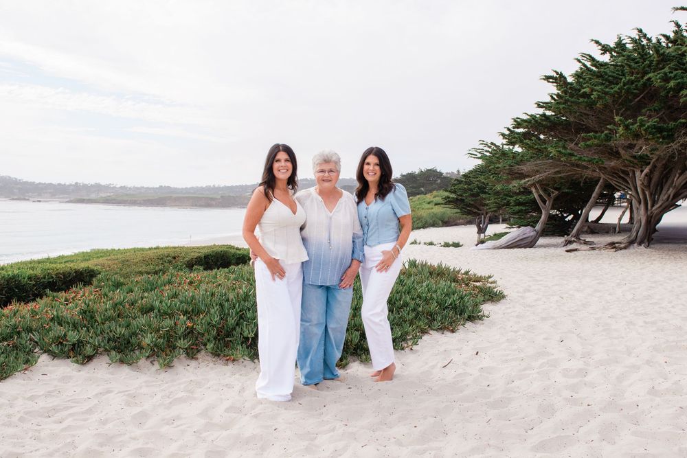 Three women in white and light blue outfits stand arm in arm on a sandy beach near coastal greenery, cypress trees, and calm ocean water.