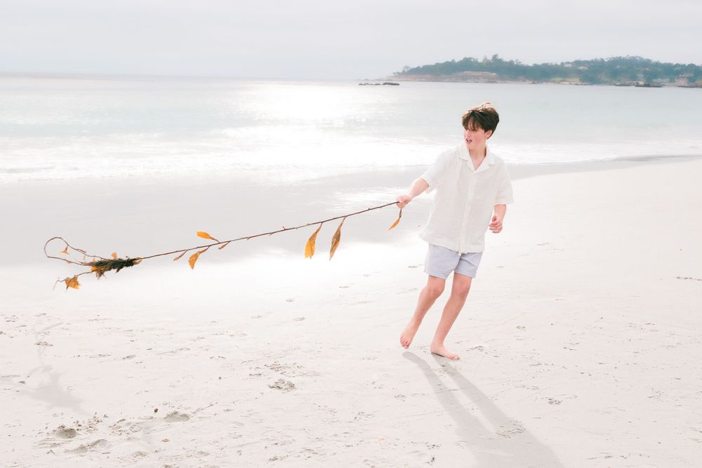 A young person in a white shirt and shorts walks barefoot on a sunny sandy beach, pulling a long strand of seaweed beside calm ocean water.