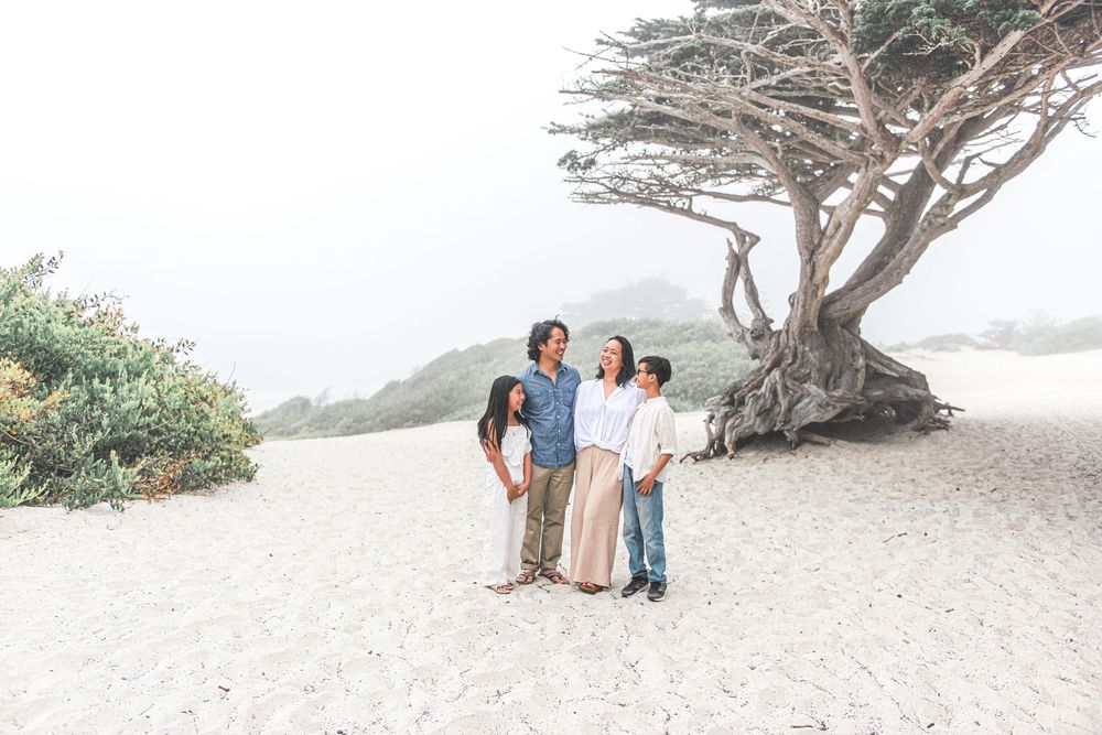 Family of four in casual clothes stands together on a white sand beach beside a windswept cypress tree on a foggy coastal day.