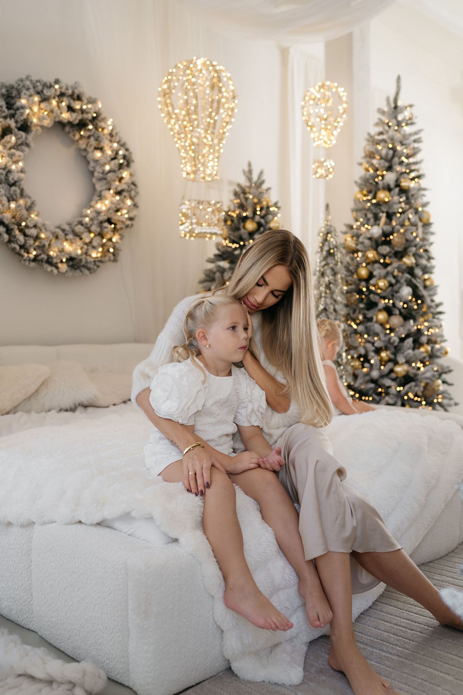 A mother and daughter in white sit on a fluffy bed with a snowy wreath, glowing light decor and frosted Christmas trees nearby.