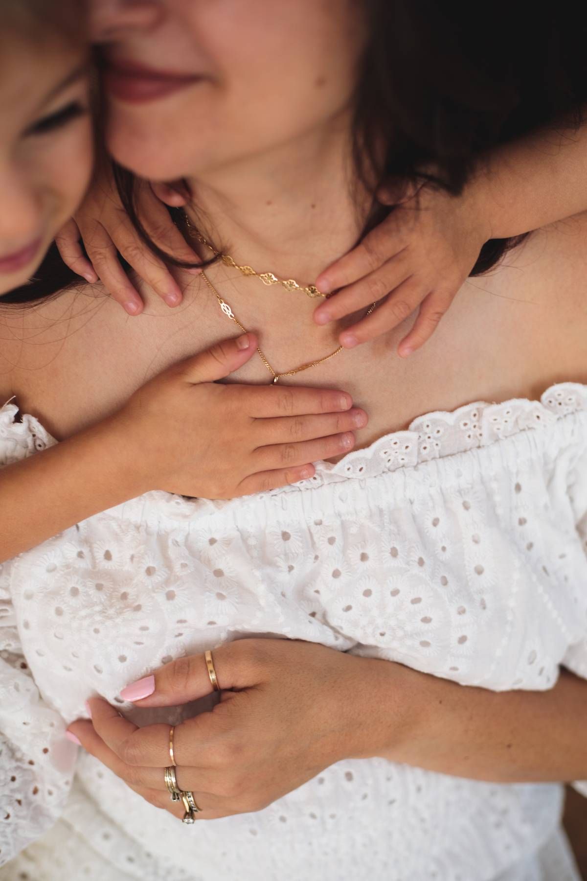 Smiling mother in a white eyelet dress is hugged by a child, with gold necklace and rings visible in a tender portrait.