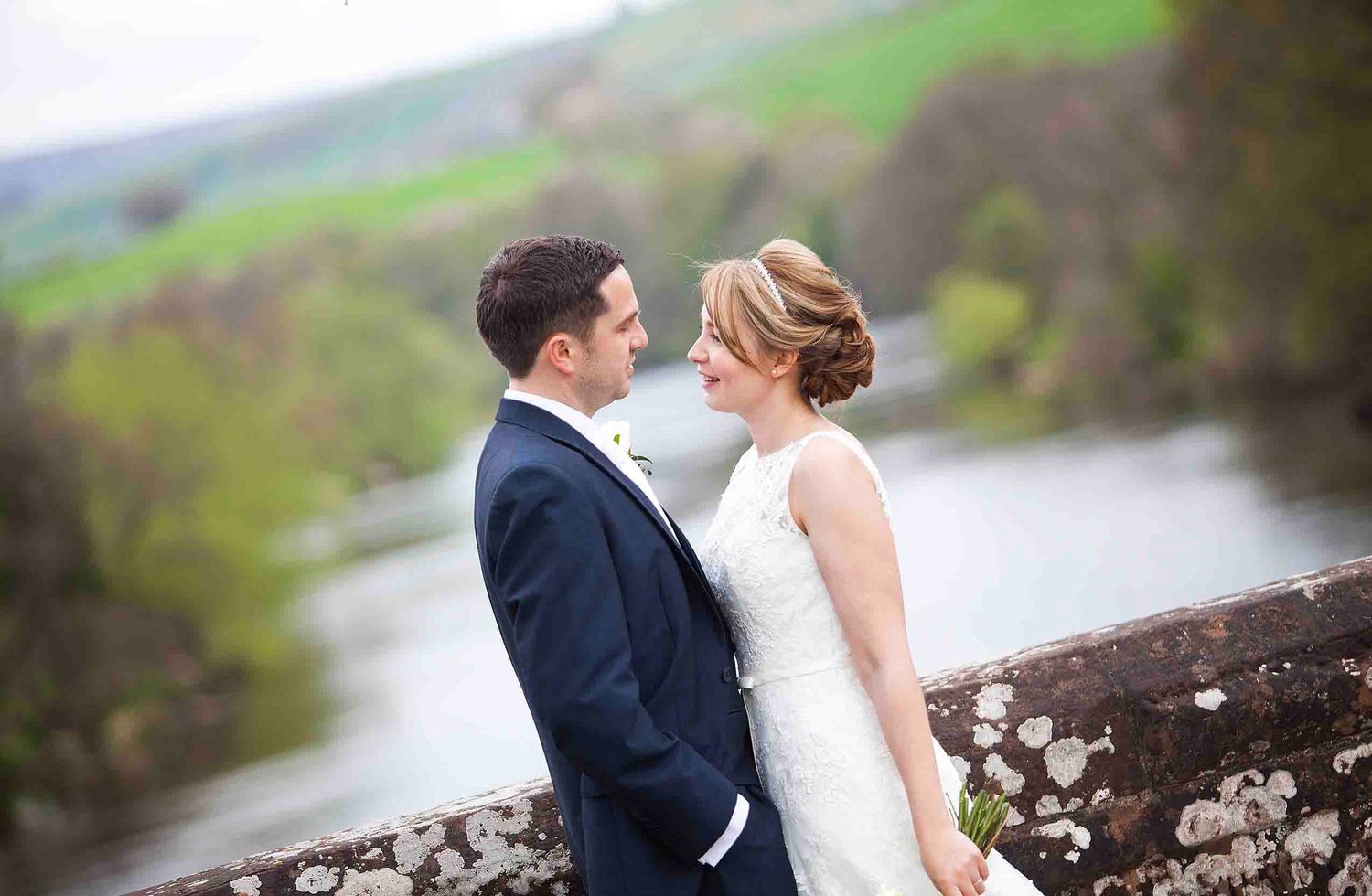 Bride in a white wedding dress and groom in a navy suit facing each other beside a stone wall, with a river and green hills behind them.