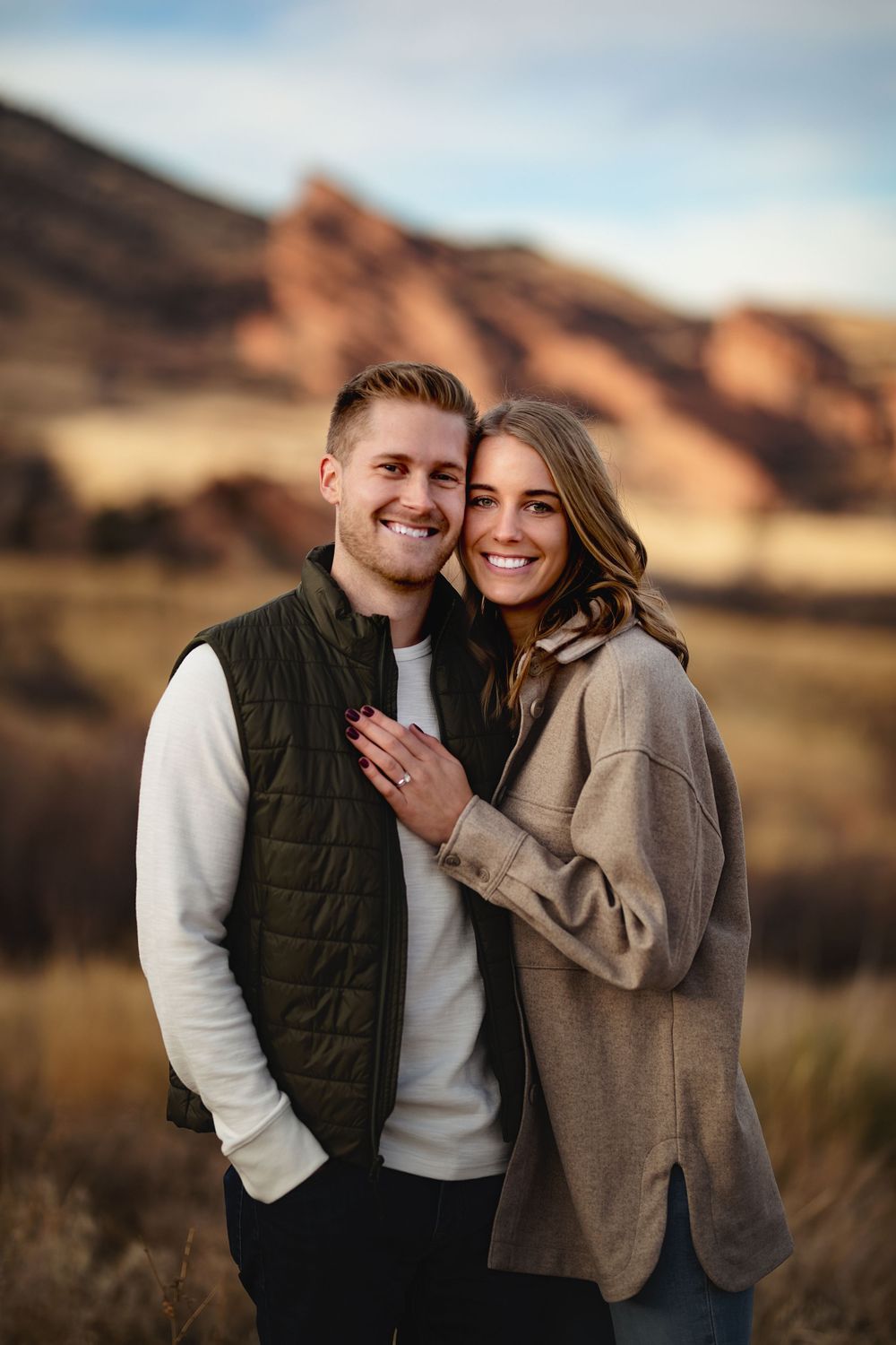 Smiling couple embracing outdoors, the woman showing her ring, with red sandstone rock formations and golden grass in the background.