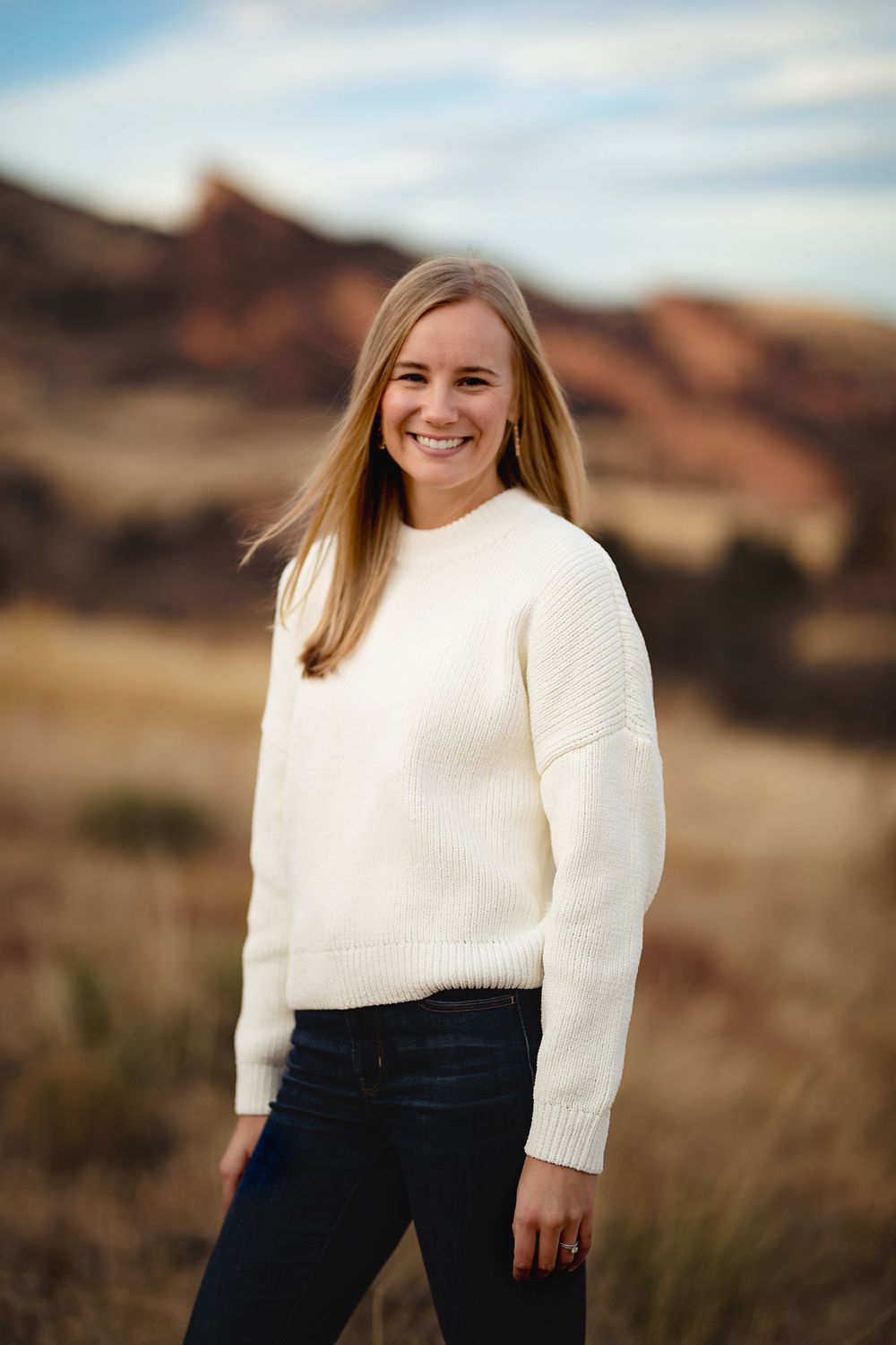 Smiling woman with long blonde hair in a cream knit sweater and dark jeans standing in a grassy field with red rock hills behind her.