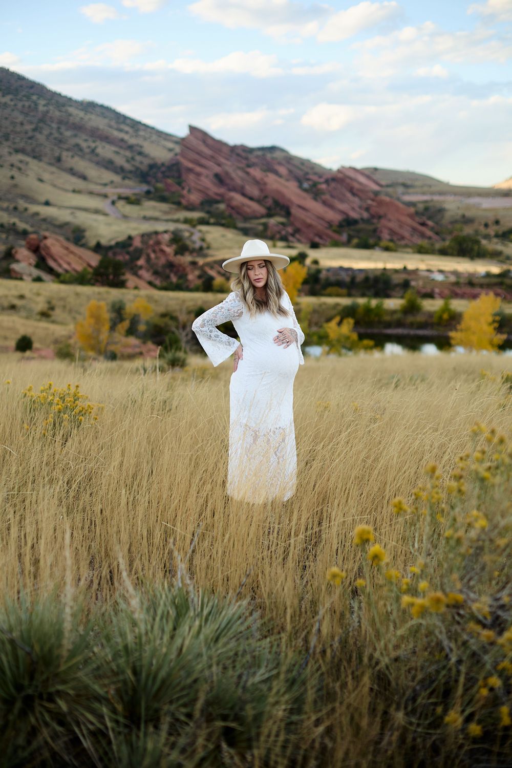 Pregnant woman in a white lace maternity dress and straw hat stands in golden grass with red rock formations and mountains behind her.