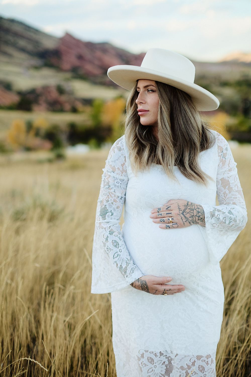 Pregnant woman in a white lace dress and wide-brim hat cradles her baby bump in a golden grass field with red rock hills behind.