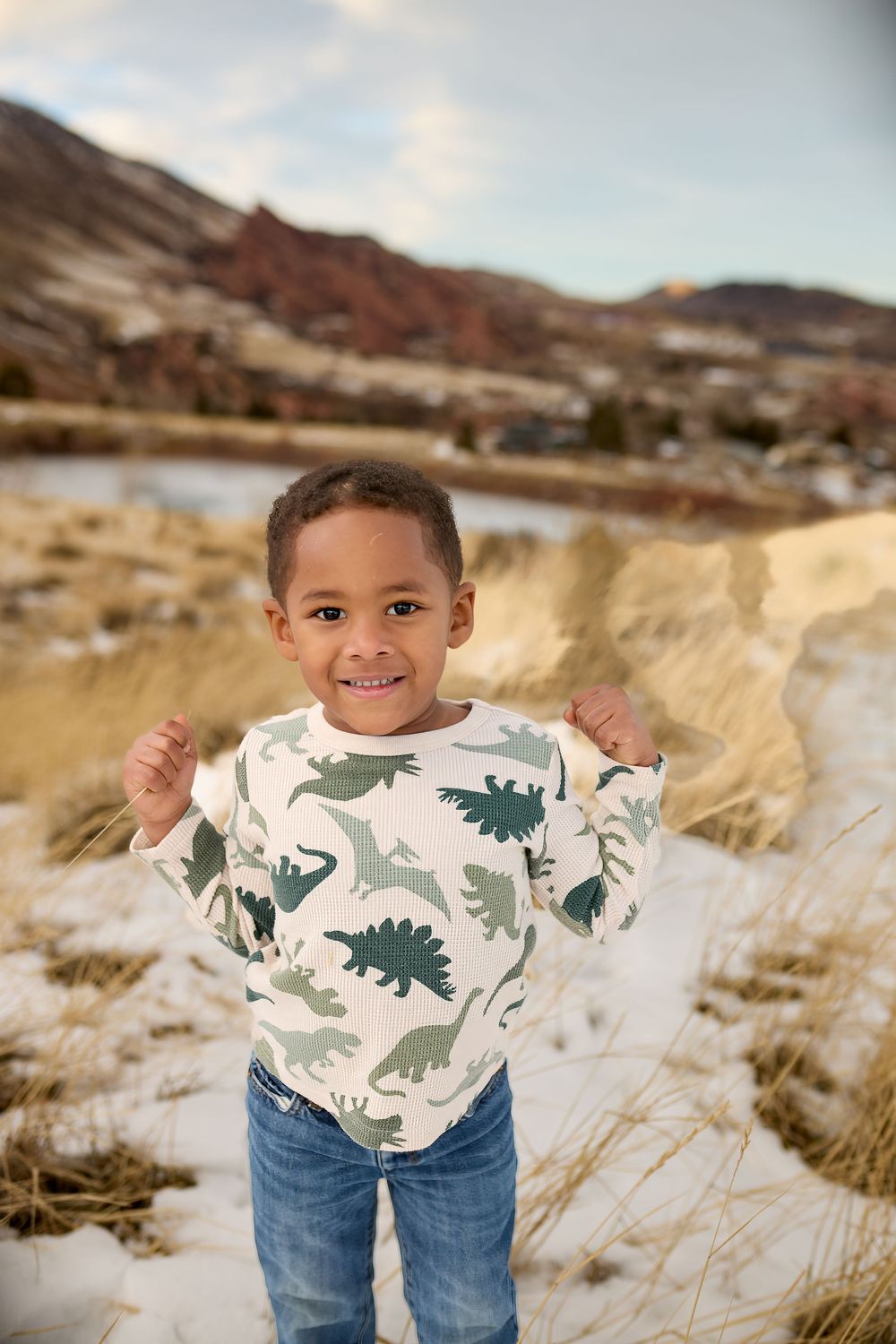 Smiling toddler in a green dinosaur-print sweatshirt and jeans stands in a snowy field with dry grass and red rock hills behind him.
