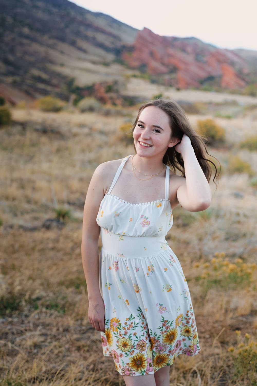 Smiling young woman in a white floral sundress touching her hair, standing in a grassy field with red rock mountains behind her.
