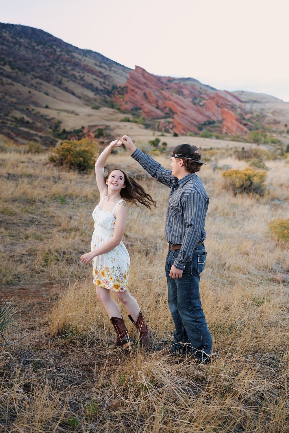 Couple dancing outdoors as a man twirls a smiling woman in a sundress and cowboy boots in a grassy field with red rock hills behind.