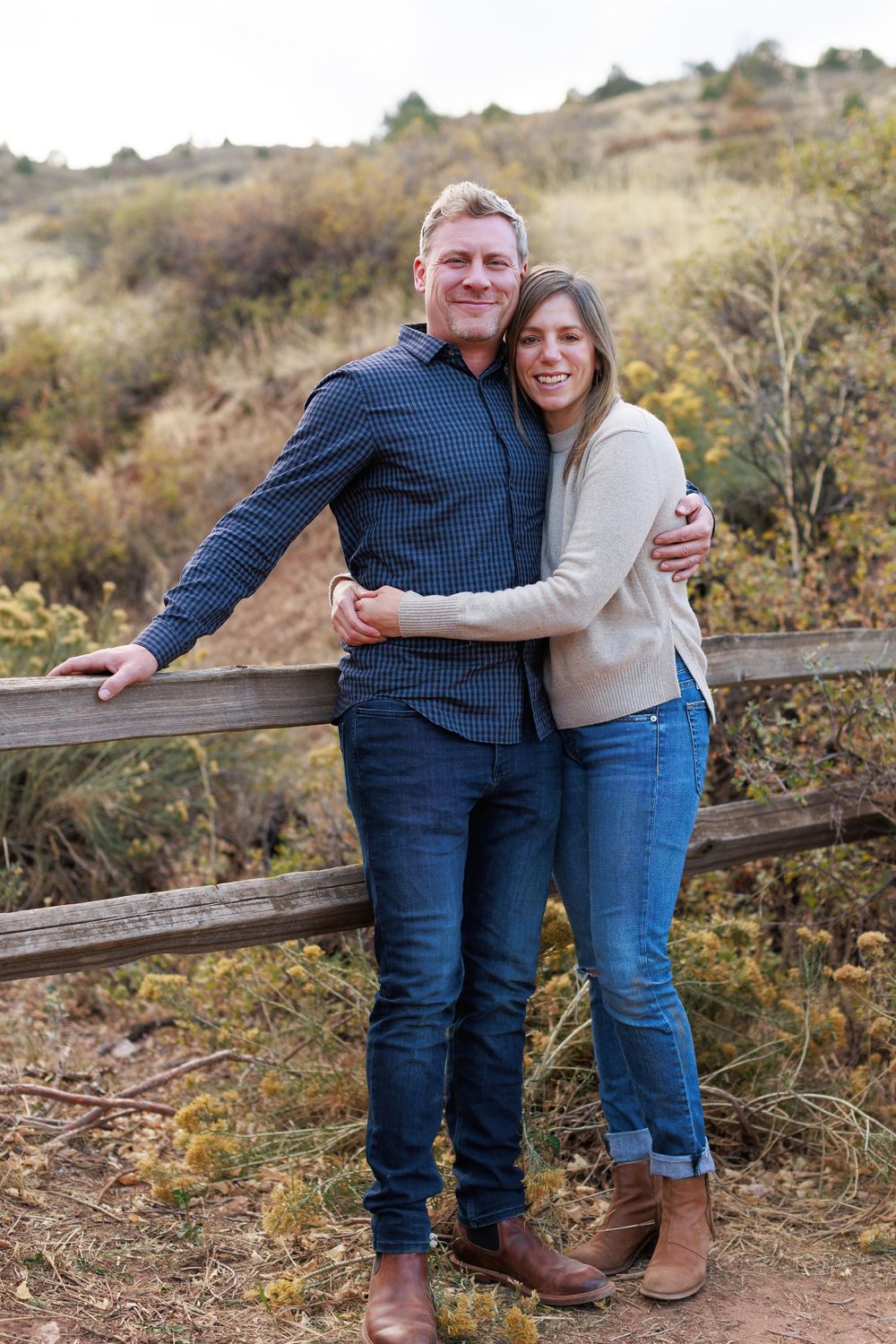 A smiling man and woman embrace beside a wooden rail fence on a sunny hillside with golden shrubs and dry grass.
