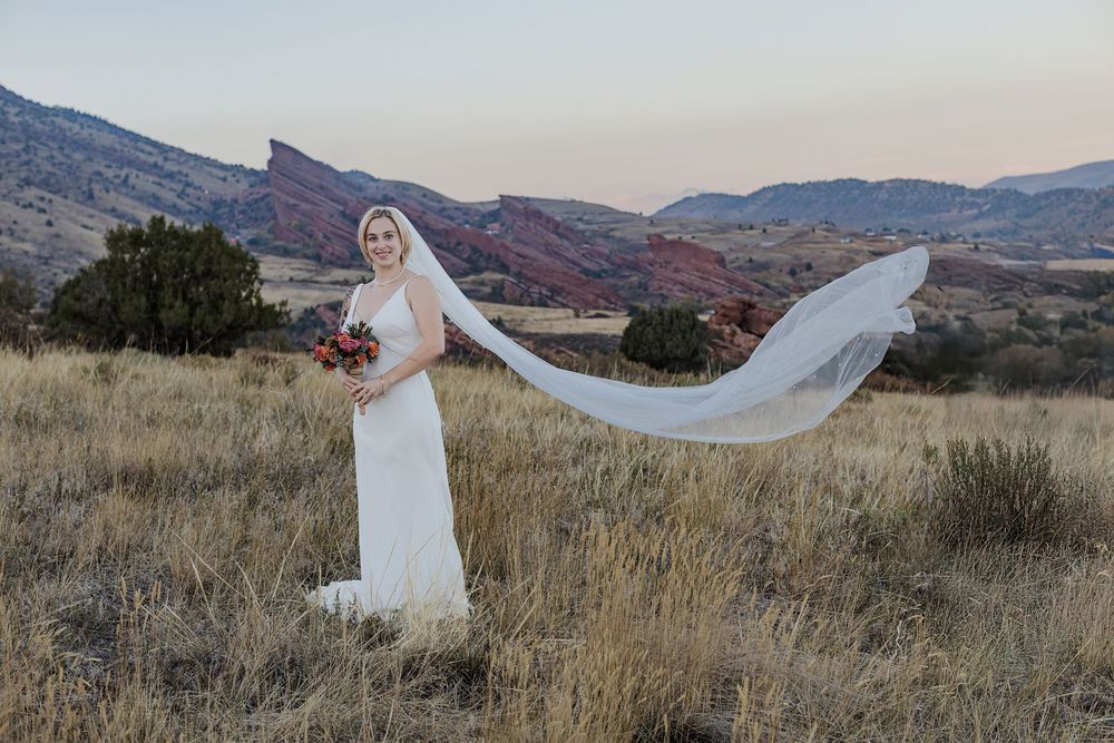 Bride in a white gown holding a bouquet as her long veil flows in the wind across a golden grass field with red rock hills at sunset.