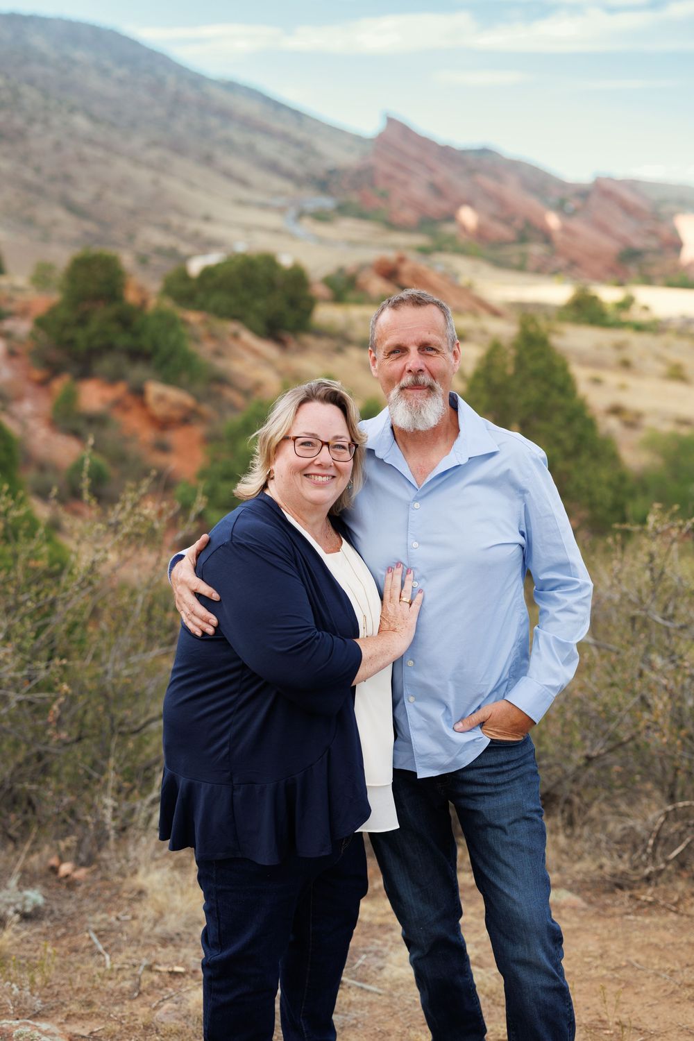 Smiling middle-aged couple embracing outdoors, with red rock formations, green shrubs, and mountains behind them at a scenic overlook.