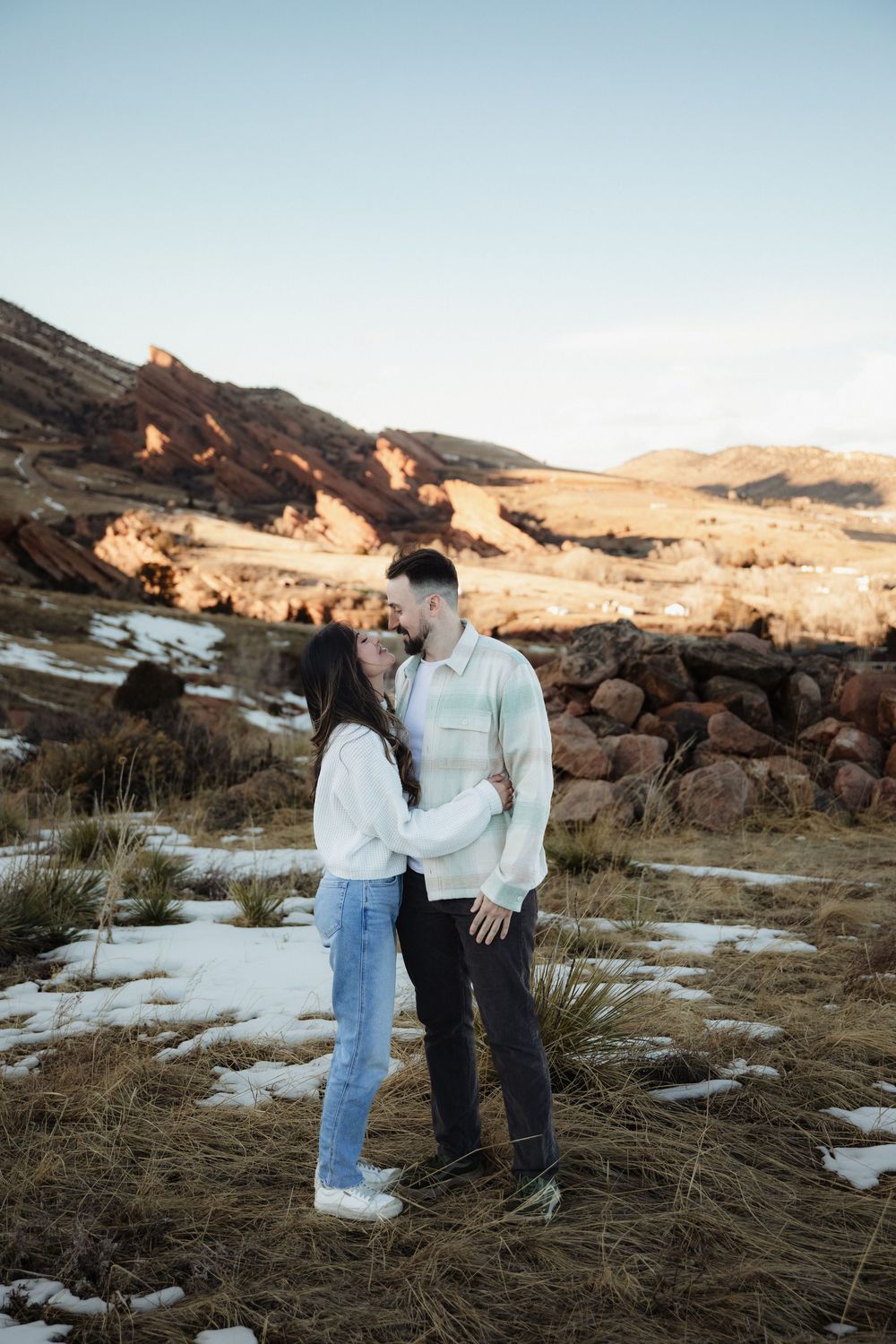 Couple embracing in a snowy field with red sandstone rock formations and mountains behind them during an engagement photo session.